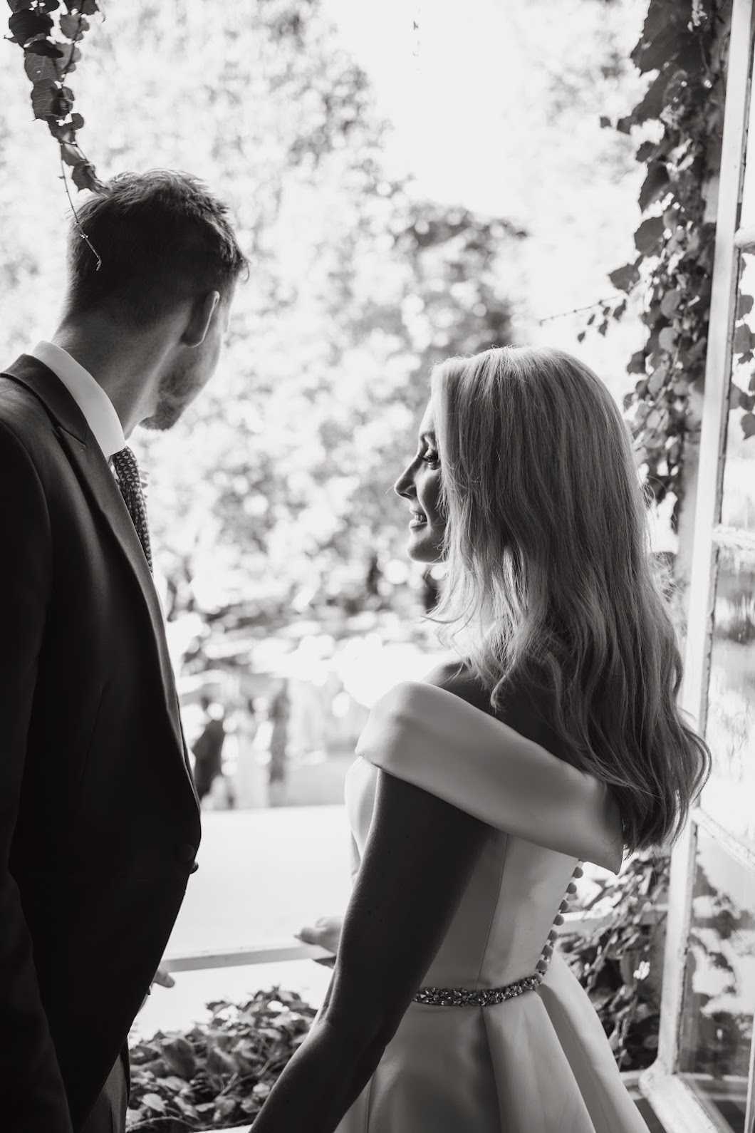 Black-and-white portrait of a bride and groom standing close together near what appears to be a doorway or window frame with climbing foliage, likely outdoors or at the threshold of an indoor-outdoor space. The bride, seen from behind at a three-quarter angle, wears an off-the-shoulder ball gown with a fitted jewelled waist belt and has long, loosely waved hair; she is smiling and looking up at the groom. The groom, also seen from behind, wears a dark suit with a patterned tie. The image is shot with high contrast, with a bright, softly bokeh'd background of foliage creating a dark-to-light tonal range that draws focus to the couple. Close-up portrait composition with shallow depth of field.