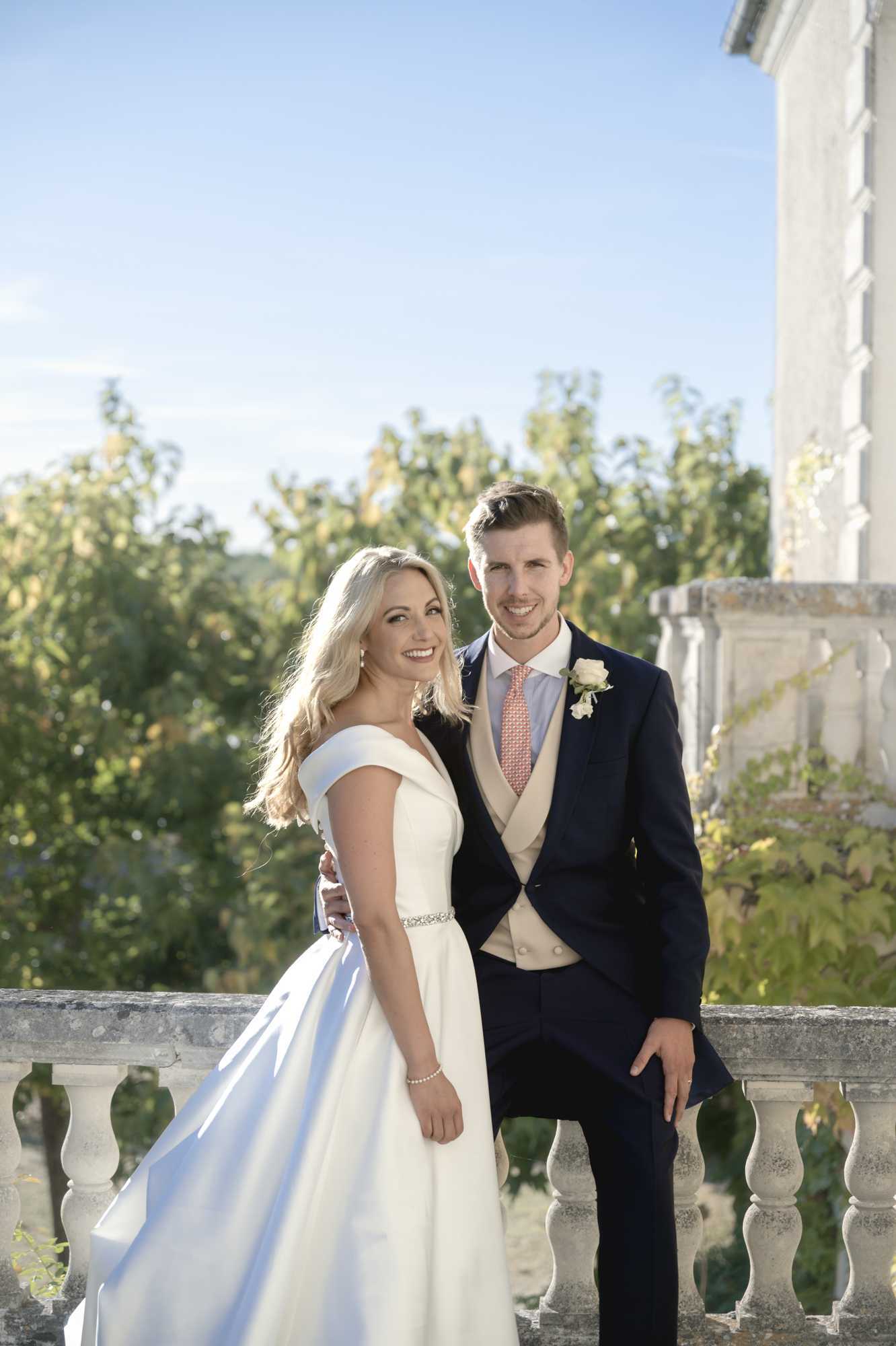 A couple poses for a portrait on a stone balustrade terrace adjacent to a classical stone building, likely a chateau or manor house. The bride wears an off-the-shoulder ivory ballgown with a slim crystal-embellished waist belt, and the groom wears a navy morning suit with a champagne waistcoat, coral patterned tie, and a white rose buttonhole. The shot is a medium portrait taken in warm natural daylight, with the bride turned slightly toward camera while the groom leans casually against the balustrade. The overall styling is classic and formal.
