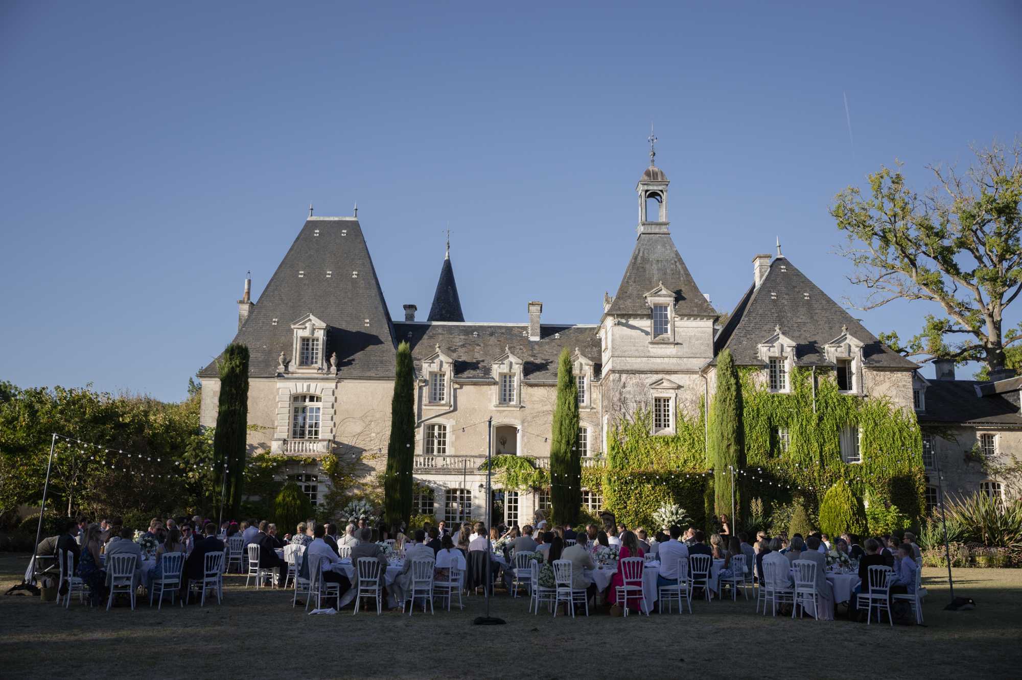 An outdoor wedding reception dinner is taking place on the lawn directly in front of a large French chateau with distinctive pointed slate roof towers, stone facade, and climbing green ivy. Approximately 80–100 guests are seated at round tables dressed in white linens, with white Chiavari chairs arranged across the grass. Floral centerpieces in white and green tones are visible on the tables. String bistro lights are strung between poles above the dining area, adding warm ambient lighting as the sun is low. The wide shot is taken from across the lawn, capturing the full scale of both the chateau architecture and the reception layout. Potential venue feature image.
