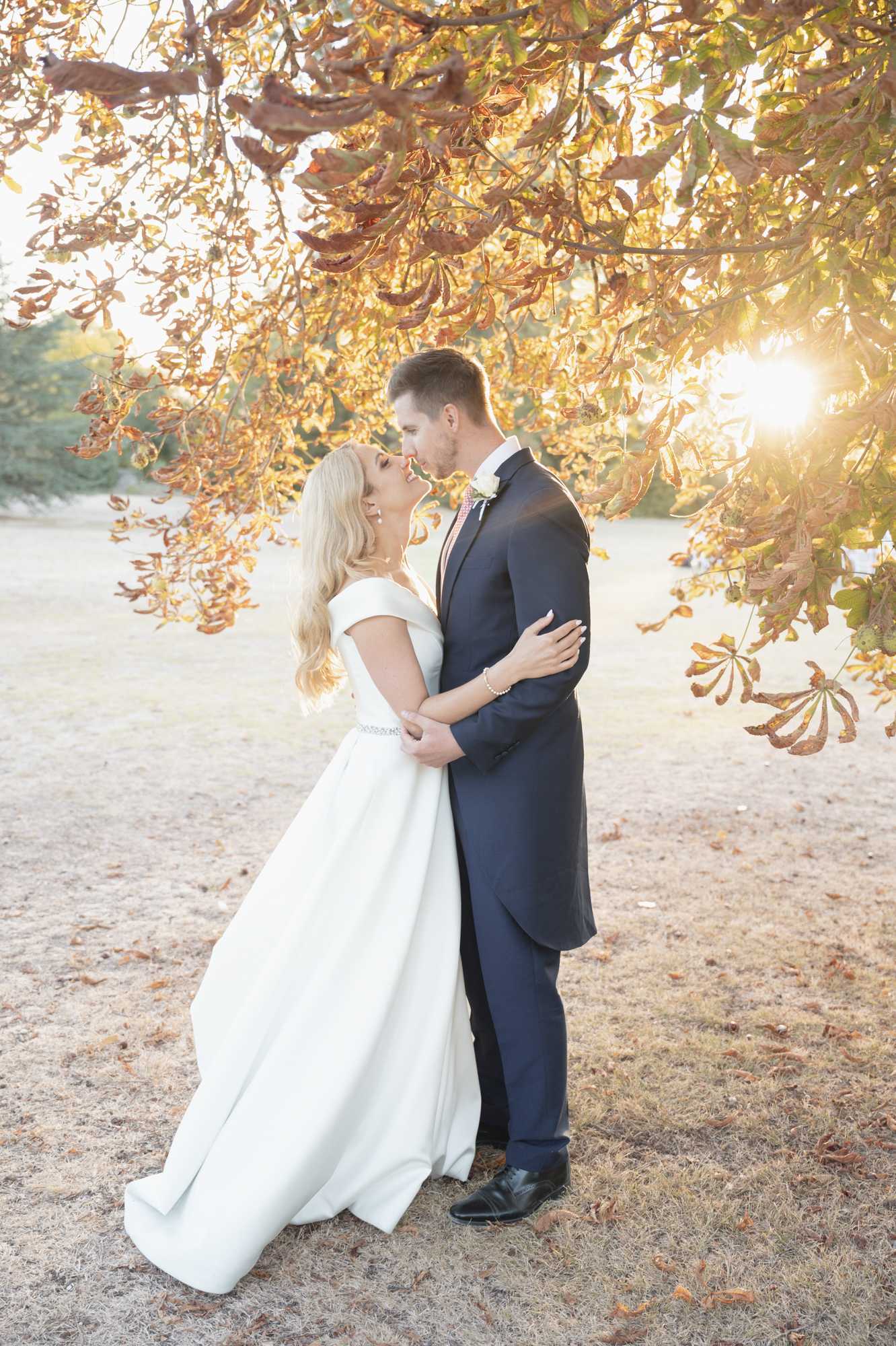 An outdoor couple portrait taken during golden hour, with the bride and groom embracing and about to kiss beneath a large tree with rust-orange and amber autumn foliage. The bride wears an off-the-shoulder ivory ball gown with a slim embellished waist belt and has long blonde hair worn down in loose waves; the groom wears a navy suit with a pink tie and a white boutonniere. Warm sunlight flares through the branches behind them, casting a golden backlit glow across the scene. The shot is a full-length portrait composition framed by the overhanging autumn canopy.