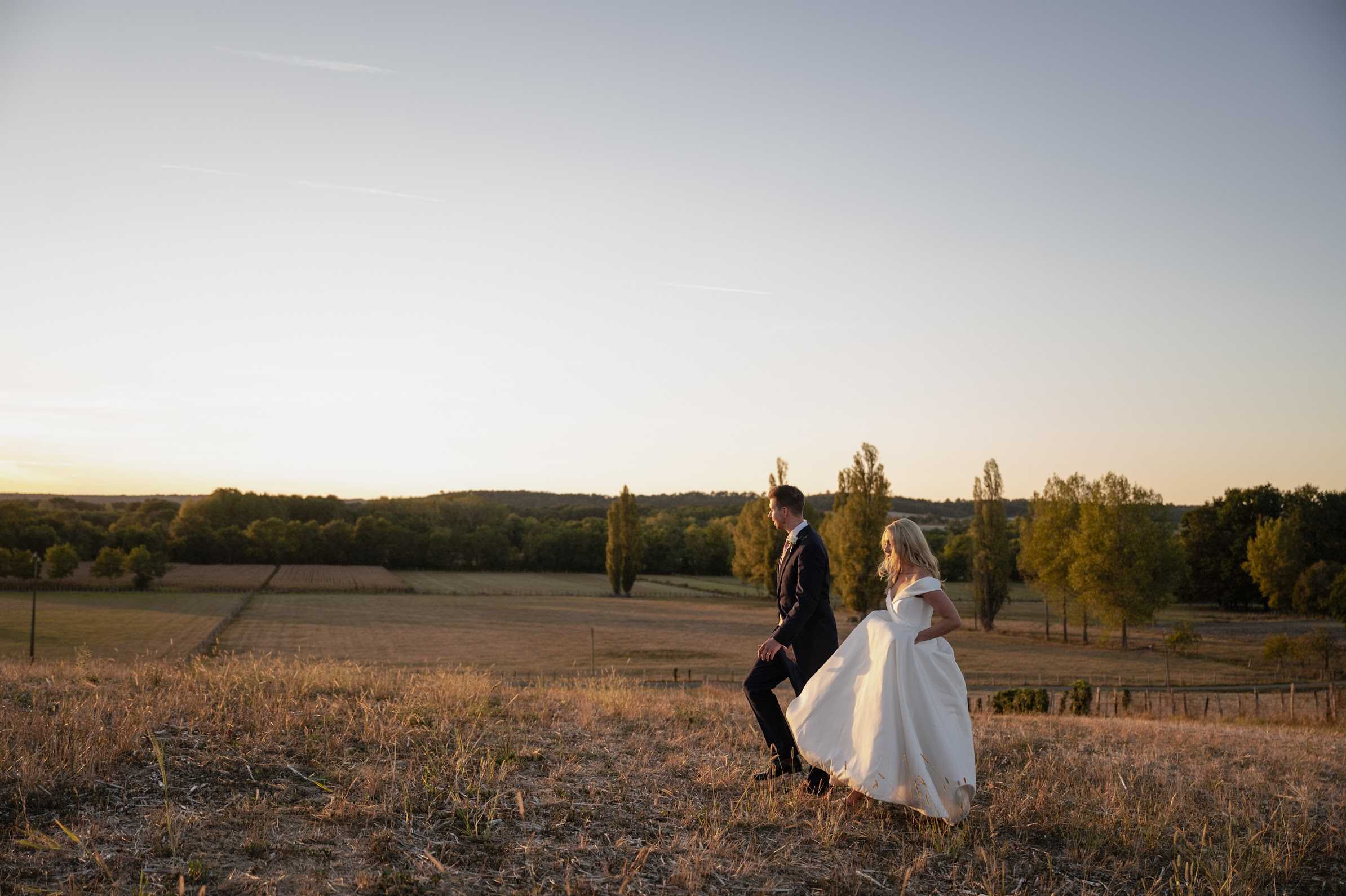 A couple portrait taken outdoors at golden hour, with the bride and groom walking through an open field with a wide rural French countryside landscape stretching behind them. The groom wears a dark navy suit and the bride wears an off-the-shoulder white ball gown with a full skirt that is in motion as she walks. The composition is a wide shot that places the couple in the lower right third of the frame, emphasizing the expansive rolling landscape, distant treeline, and pale golden sky at sunset. The warm late-day light bathes the scene in amber tones.