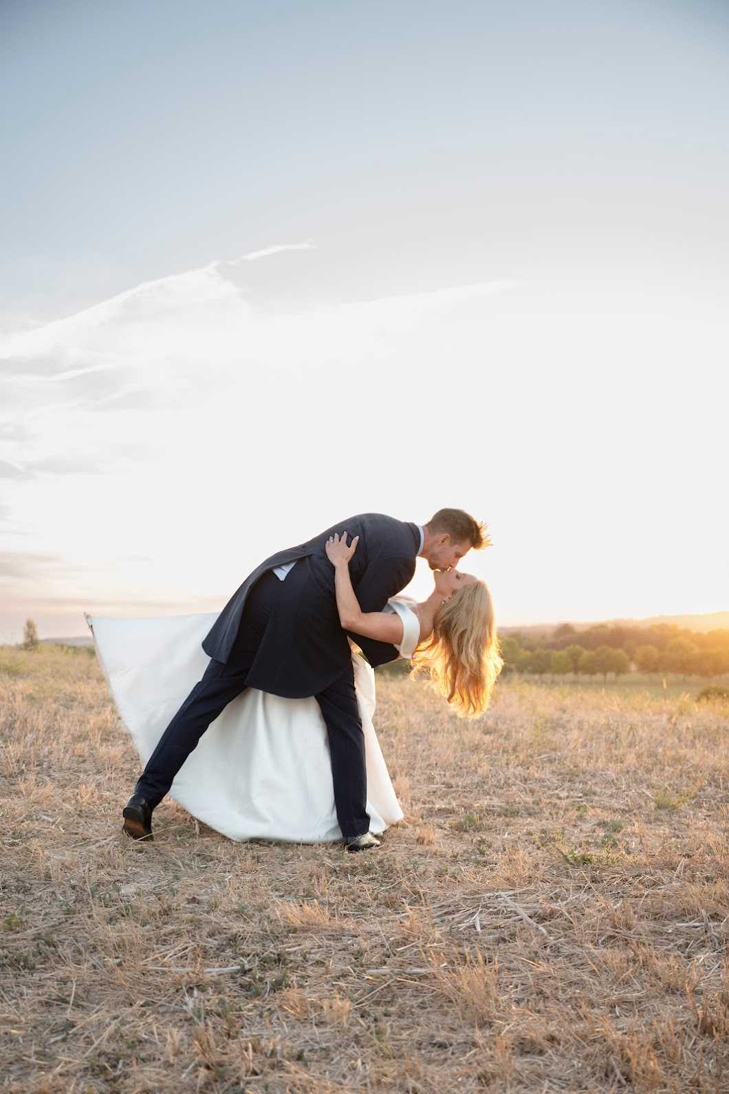 A couple portrait taken outdoors in an open field during golden hour, with warm sunset light backlit behind them. The groom, wearing a dark navy suit, is dipping the bride into a kiss while her long cathedral-length veil billows out to the left in the wind. The bride wears a white gown with a full skirt and has long blonde hair. The composition is a full-length wide shot with the couple centered against a wide open landscape and a pale evening sky.