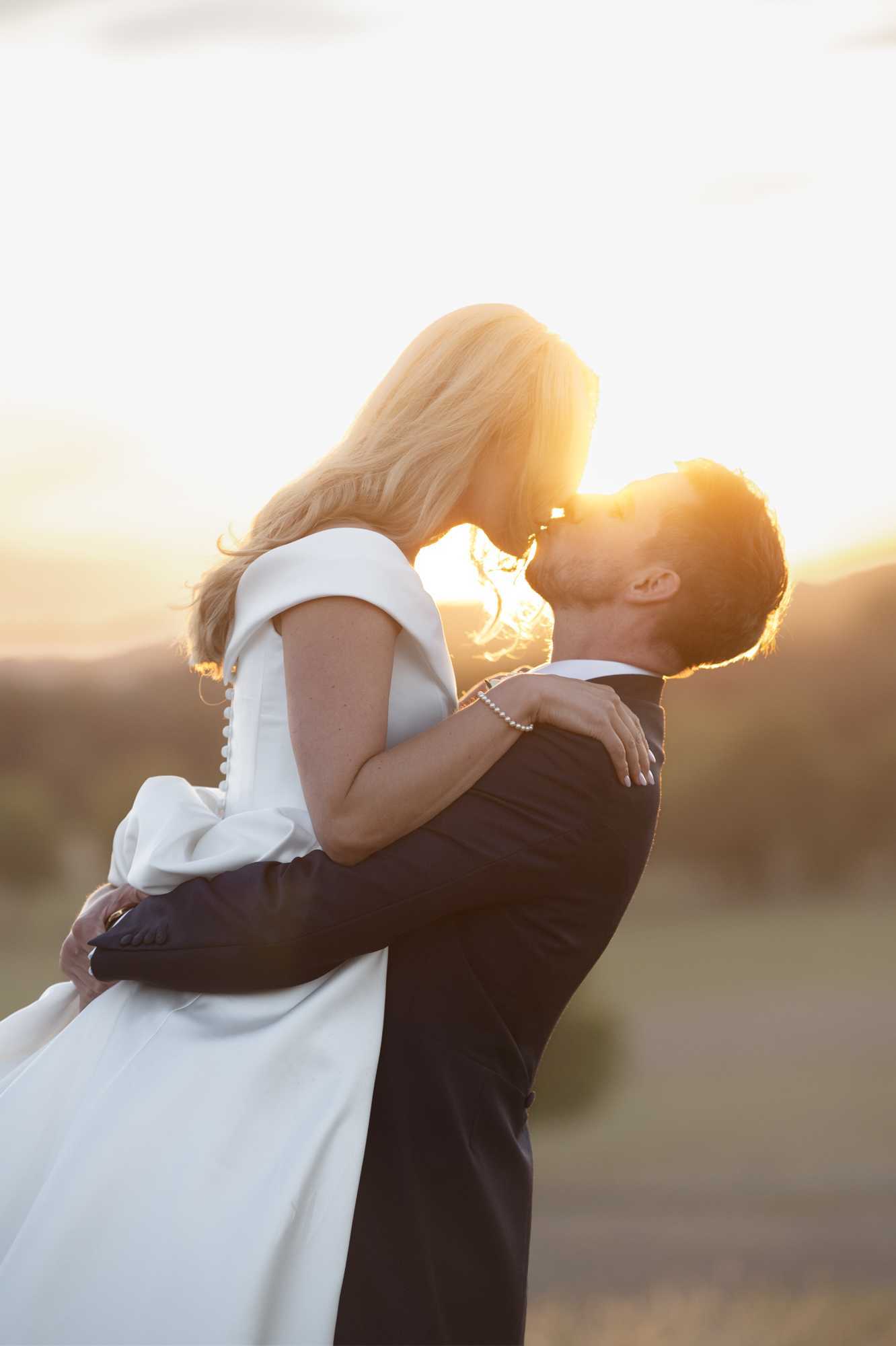 A couple portrait shot outdoors during golden hour, with the groom lifting and kissing the bride against a warm backlit setting. The bride wears a white off-the-shoulder gown with a clean, modern silhouette and button-back detail, paired with a pearl bracelet, and has long blonde hair. The groom is dressed in a dark navy or black suit with a white dress shirt. The background is heavily blurred with warm golden tones suggesting an open landscape or countryside setting. The composition is a mid-shot portrait with strong backlighting creating a glowing rim-light effect around the couple.