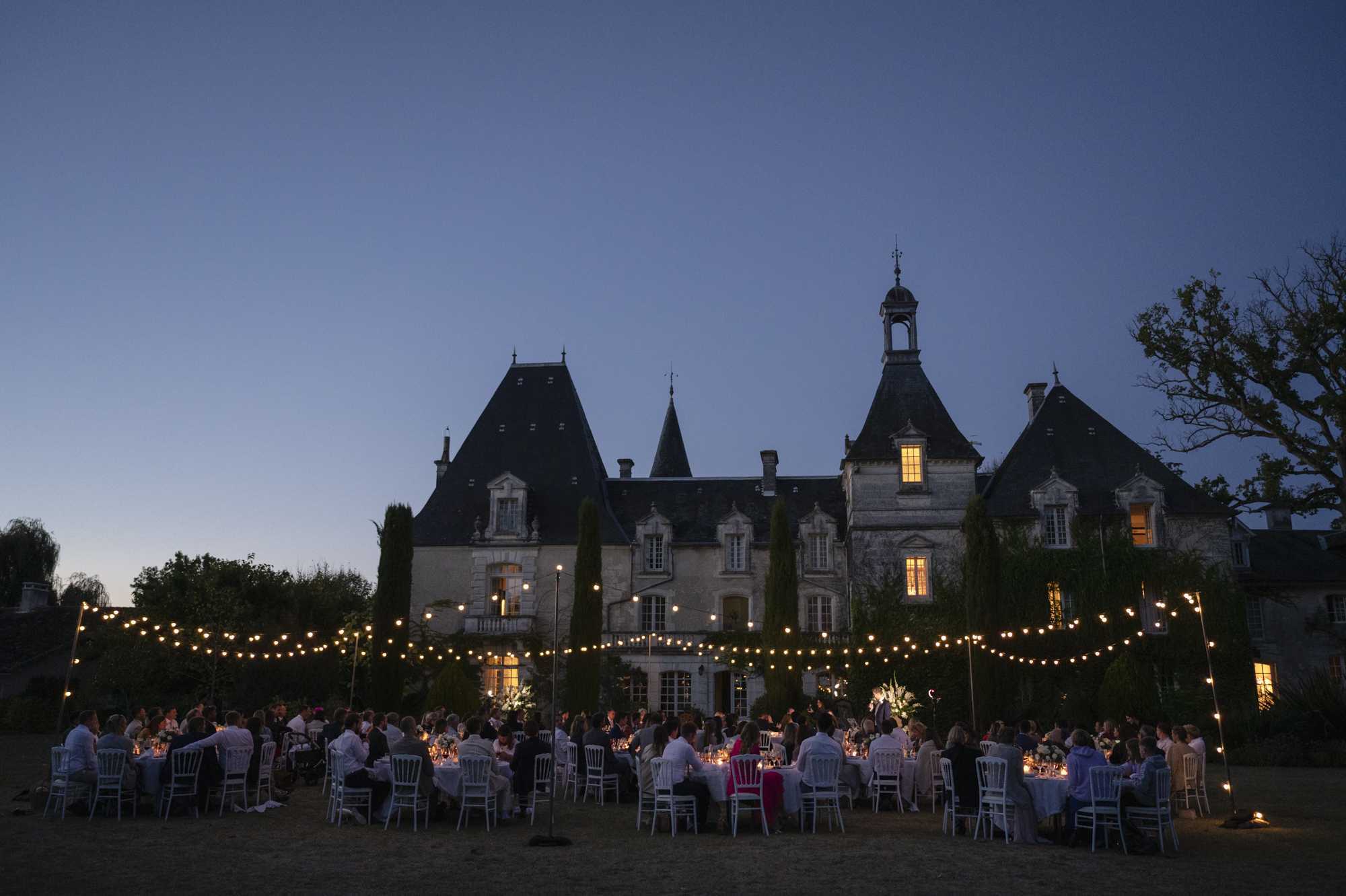 An outdoor wedding reception dinner is taking place on the grounds of a French château at dusk, with approximately 80–100 guests seated at round tables covered in white linens and arranged with white Chiavari chairs. Multiple strands of warm Edison bulb fairy lights are strung on tall poles across the lawn in front of the château, providing the primary decorative lighting as natural light fades. The château itself is a classic French manor with steep slate-roofed turrets, stone façade, and ivy-covered walls, its windows glowing warmly from interior lighting. The wide-angle shot captures the full scale of the reception setup against the silhouette of the building at twilight, emphasizing the relationship between the outdoor dinner setting and the architectural backdrop. Potential venue feature image.