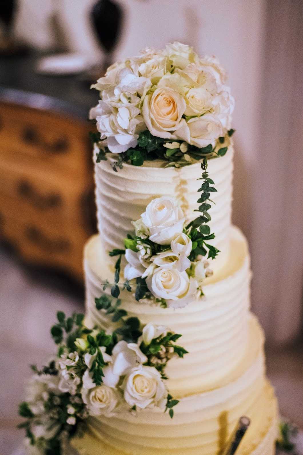 Close-up detail shot of a three-tier wedding cake with ivory buttercream frosting featuring a horizontal textured ruffled finish on each tier. The cake is decorated with a cascading arrangement of cream roses, white hydrangeas, white freesias, and green eucalyptus trailing diagonally from the top tier down to the base. The top is crowned with a dense cluster of cream roses and white hydrangeas. The background is softly blurred, revealing what appears to be a wooden antique dresser, suggesting an indoor reception setting with a classic decor style.