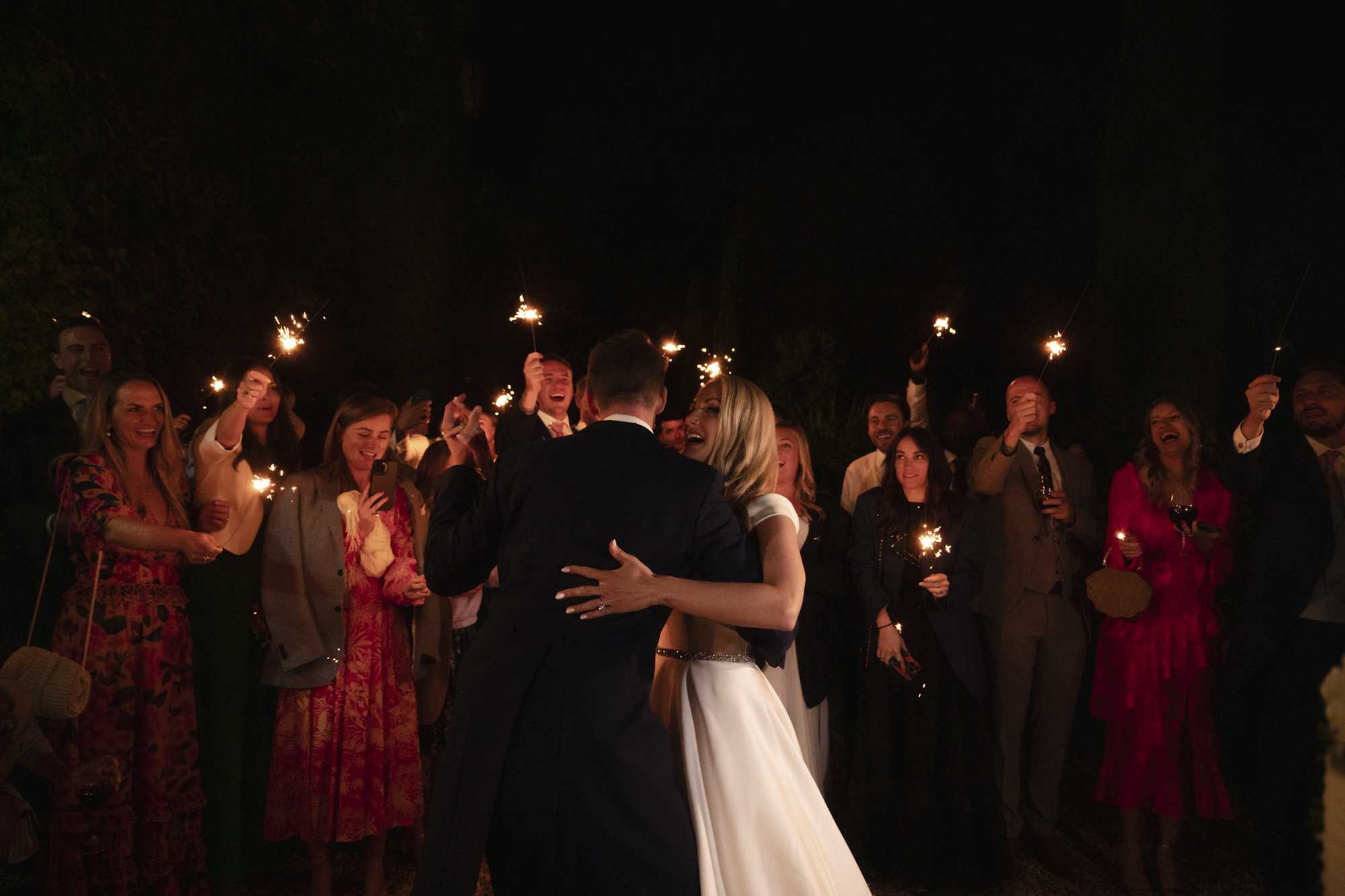 The couple shares a first dance outdoors at night, surrounded by approximately 20 guests holding lit sparklers that illuminate the scene with warm golden light. The bride wears a sleek white gown with a beaded waistband detail and cap sleeves, while the groom wears a navy suit; she is laughing and facing the camera as they embrace. Guests are dressed in a mix of formal attire including a red floral maxi dress, a fuchsia dress, and dark suits, many smiling and cheering while holding their sparklers aloft. The wide shot captures the couple centered in the frame with guests forming a loose circle around them against a dark night-sky background.