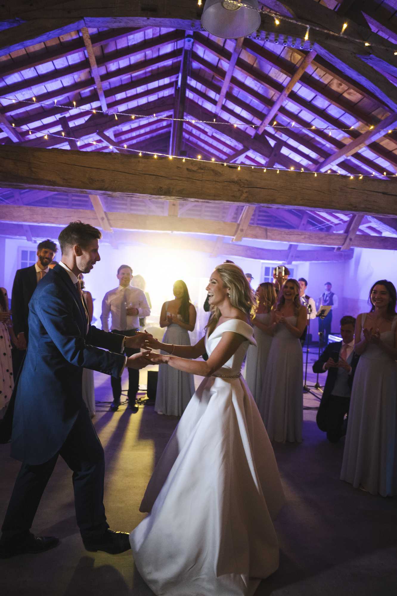 The couple's first dance is taking place inside a rustic barn venue with exposed wooden beam ceiling rafters strung with warm fairy lights and lit by purple uplighting. The groom wears a deep navy blue suit with a pink tie, and the bride wears an off-the-shoulder ivory satin ball gown with a fitted waistband detail; both are holding hands and laughing. Approximately ten to fifteen guests encircle the dance floor watching and clapping, with bridesmaids in floor-length grey gowns visible on the right side and a live band performing in the background. The image is a wide portrait-orientation shot taken from a low angle, capturing the full scene with dramatic purple and warm amber stage lighting creating a lively reception atmosphere.