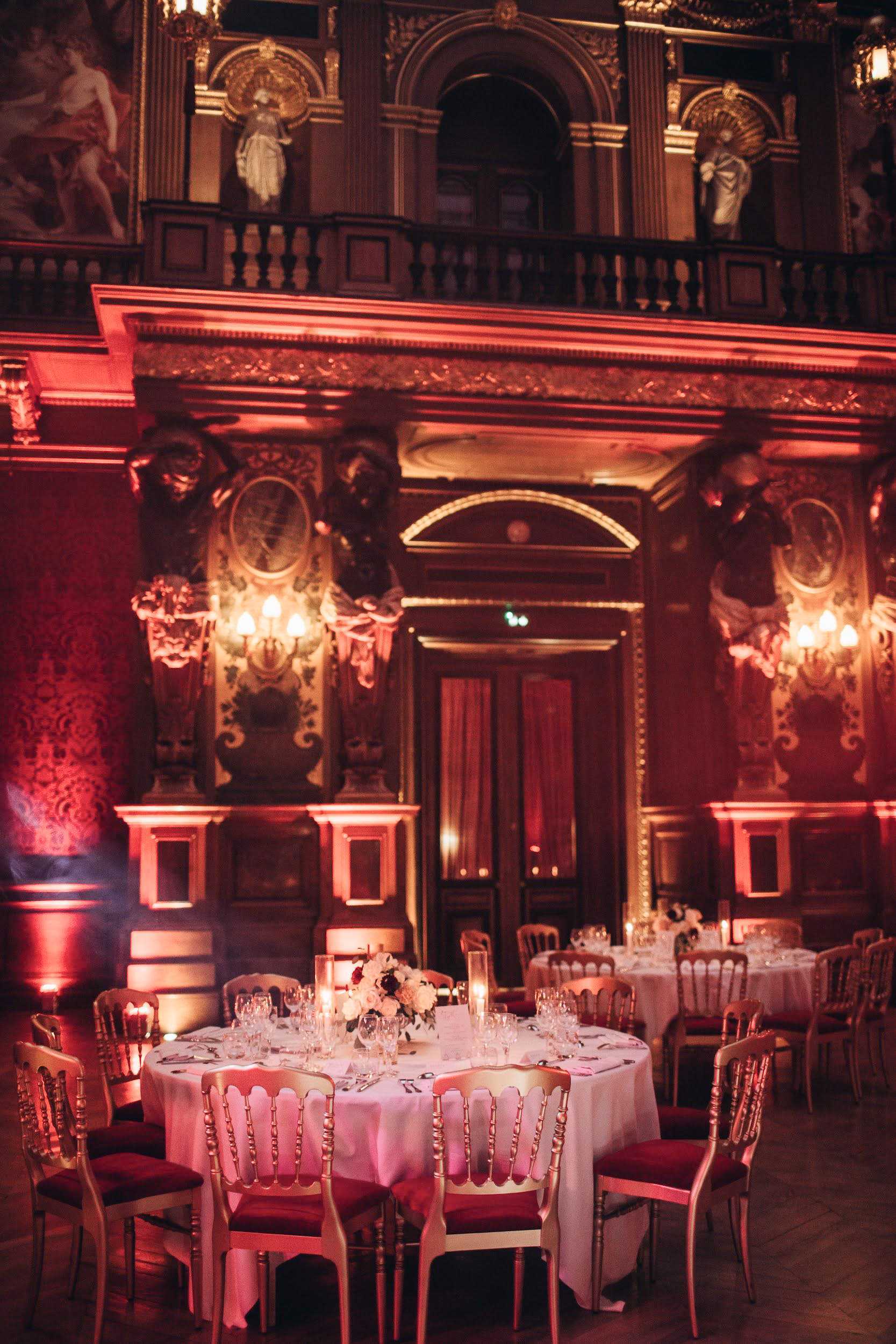 A wedding reception setup inside a grand baroque-style ballroom, bathed in deep red and amber uplighting that illuminates the ornate gilded architectural details, sculpted figures on an upper balcony, wall-mounted candelabras, and heavily decorated pilasters. Round tables are dressed in white linen and set with crystal glassware, with low centerpieces of ivory and burgundy flowers with dark foliage, surrounded by gold Napoleon-style chairs with deep red upholstered seats. Candles in tall glass holders are placed on each table, adding warm ambient light. No guests are present in the shot, which is a wide-angle interior view taken at mid-height. Potential venue feature image.