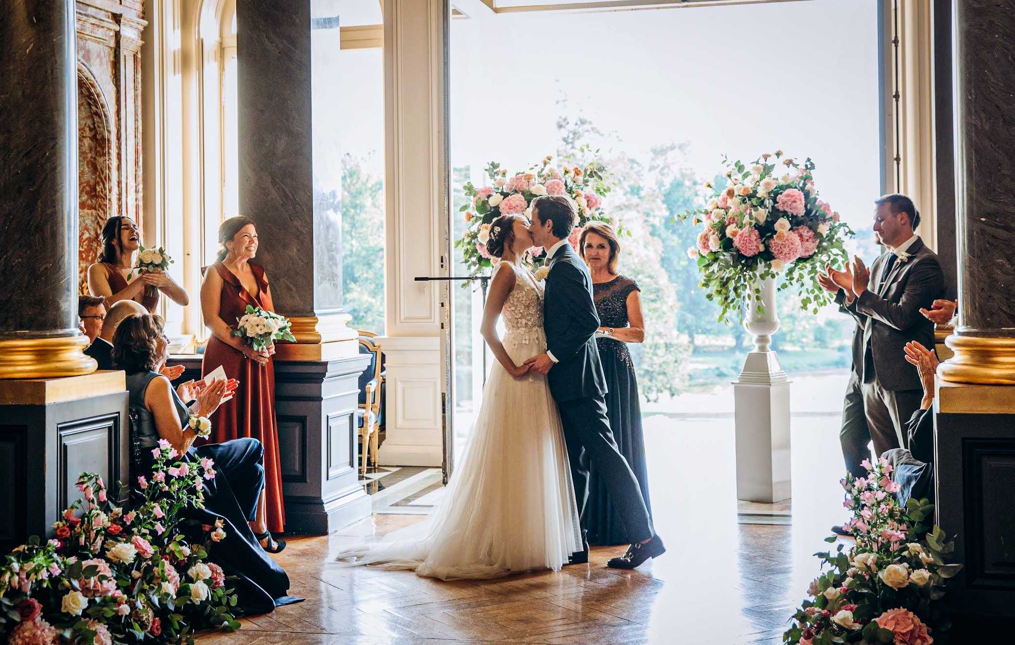 The bride and groom share their first kiss during an indoor ceremony inside what appears to be a grand chateau or palace salon, with tall ornate columns with gold detailing and large open doors revealing a garden beyond. The bride wears a white tulle ballgown with floral appliqué detailing on the bodice, while the groom is in a dark navy suit. Two large white pedestal urns flank the couple, each holding oversized arrangements of blush pink peonies, white roses, and eucalyptus; matching low floral arrangements of pink, blush, and white blooms line the aisle floor on both sides. Approximately six guests and two bridesmaids in rust-red wrap dresses are visible reacting with applause and laughter, along with a woman in a dark beaded gown standing nearby, and a photographer kneeling in the aisle at lower left. The wide-angle shot captures the full ceremony space, the warm parquet flooring, and the natural backlight streaming through the open doors, creating a classic, formal aesthetic.