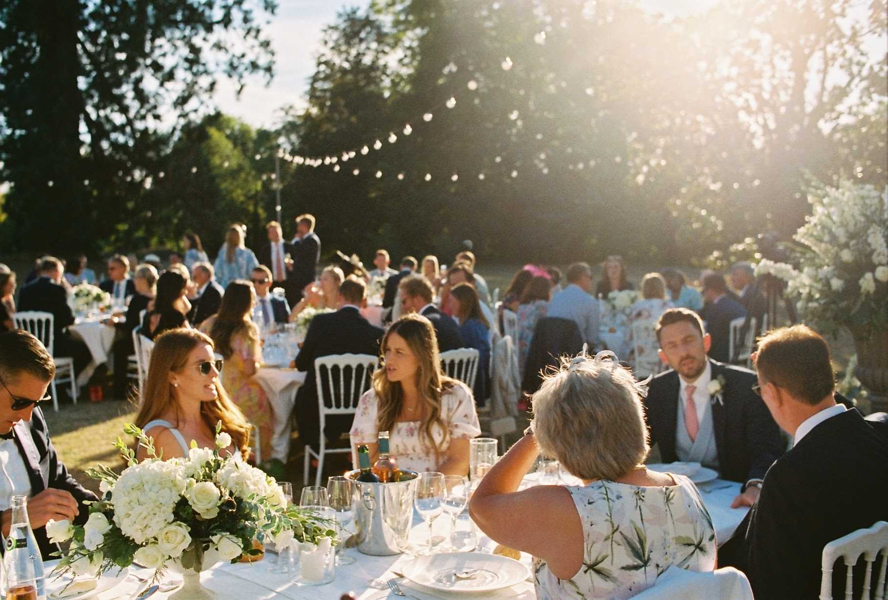 An outdoor wedding reception dinner captured during golden hour, with approximately 60-80 guests seated at multiple round tables covered in white linen. The foreground table features a centerpiece of white hydrangeas and white roses with green foliage, alongside glassware, white dinnerware, and bottles of sparkling water. Guests are dressed in a mix of dark suits with salmon-pink ties, floral summer dresses, and smart casual attire. White Chiavari chairs are used throughout. String bistro lights are strung between trees overhead, and large white floral arrangements are visible in the background right. The setting appears to be a garden or parkland, with warm late-afternoon sun creating strong lens flare across the upper right of the frame. The shot is a wide environmental portrait taken at table level, with the background slightly out of focus.