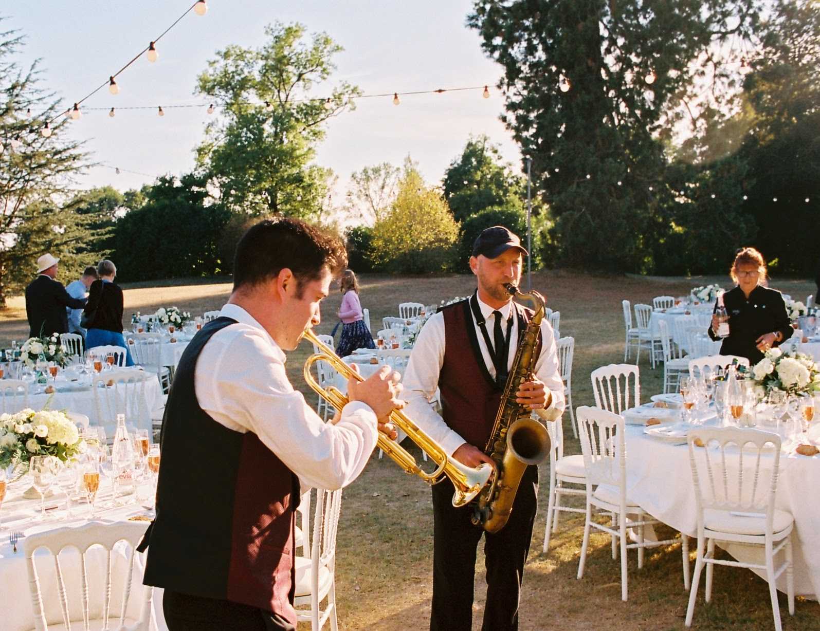Two live musicians perform during an outdoor wedding reception, with one playing a brass trumpet and the other playing a tenor saxophone; both wear matching dark burgundy vests, white shirts, and black ties. The setting is a garden or estate grounds with multiple round and rectangular tables dressed in white linen, white Chiavari chairs, and low centerpieces of white florals including what appear to be hydrangeas and roses with green foliage. Warm-toned festoon bulb lights are strung overhead, and champagne flutes are set at each place setting, catching the golden late-afternoon light. The wide shot captures roughly a dozen guests mingling in the background as the reception space is in use, with a classic, garden-party styling aesthetic shot on film giving the image a warm, slightly grainy quality.