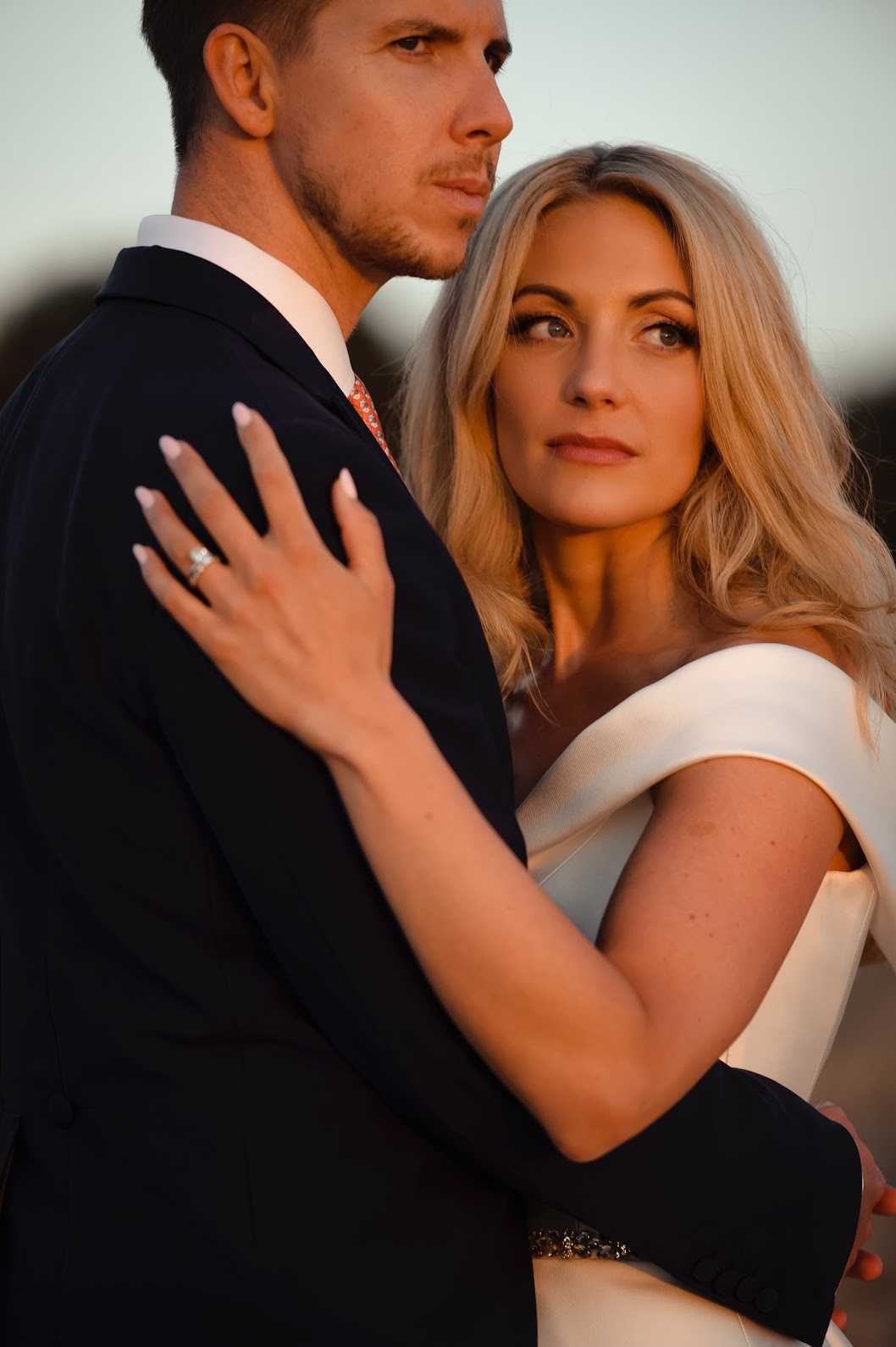 A close-up couple portrait taken outdoors during golden hour, with warm amber light illuminating the bride's face and skin. The bride wears a white off-the-shoulder gown with a clean, structured neckline and a jeweled belt at the waist, her blonde wavy hair loose, and a solitaire diamond engagement ring visible as she rests her hand on the groom's shoulder. The groom wears a navy suit jacket with a white shirt and a coral patterned tie. The composition is a tight mid-shot focused on the bride's face and the couple's embrace, with the background softly blurred and out of focus.