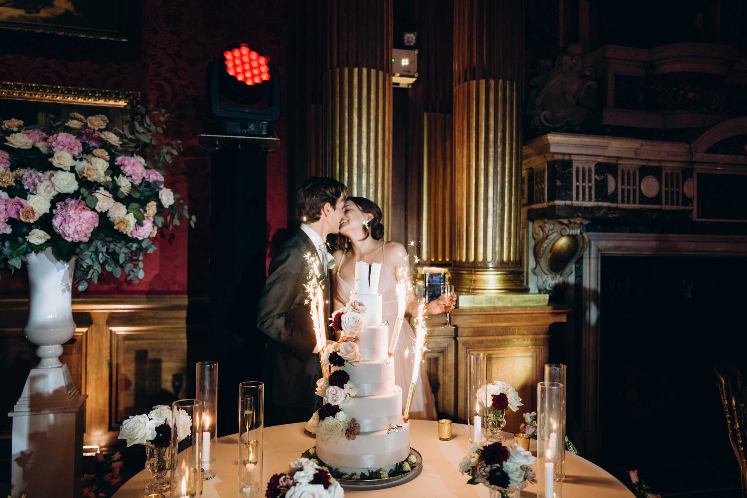 The bride and groom share a kiss during the cake cutting moment at an indoor evening reception inside a grand ballroom or stateroom featuring gilt columns, ornate wood paneling, and deep crimson walls hung with gold-framed artwork. The five-tier white fondant wedding cake is adorned with blush, ivory, and burgundy roses and is flanked by lit sparklers. The cake table is decorated with tall hurricane candle holders, low floral clusters in white, blush, and deep burgundy tones, and champagne flutes. The bride wears a simple ivory spaghetti-strap gown and the groom a grey suit. To the left, a large white urn holds an oversized arrangement of blush peonies, ivory roses, and eucalyptus. The overall decor palette is classic and rich with deep reds and golds, accented by candlelight and a visible red event lighting fixture overhead. Medium wide-angle portrait shot.