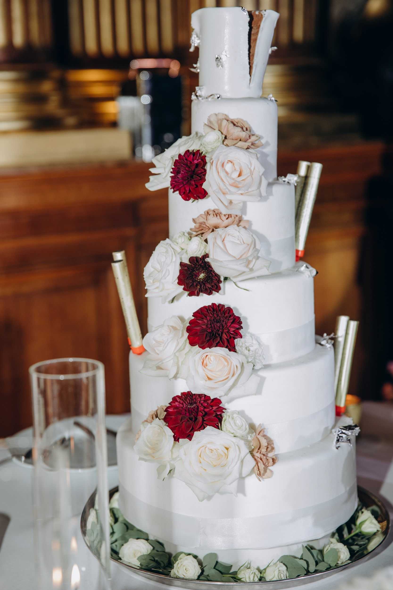 Close-up detail shot of a five-tier white fondant wedding cake displayed on a silver cake stand, set against a warm, blurred indoor background with gold architectural columns suggesting a formal ballroom or chateau reception hall. The cake is decorated with a cascading arrangement of fresh flowers including white roses, blush roses, and deep burgundy dahlias, with eucalyptus and small white spray roses at the base. Gold sparkler candles with orange tips are inserted into multiple tiers around the cake, and the top tier features a partially open section revealing a chocolate interior with silver leaf detailing. A clear glass and a lit votive candle are visible to the left of the cake stand.
