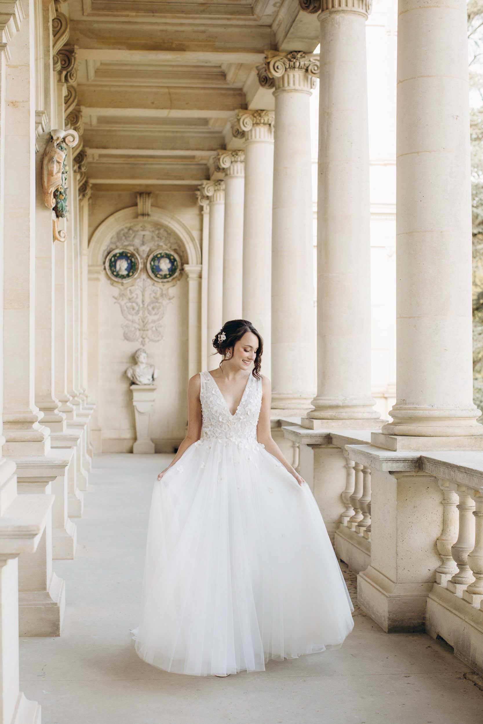 A bridal portrait taken outdoors along a grand classical colonnade, likely at a French chateau or palace, featuring tall Corinthian columns, a decorative arched niche with a marble bust and ornamental medallions, and a stone balustrade. The bride stands alone in the center of the walkway, wearing a white ball gown with a deep V-neckline, a floral appliqué lace bodice, and a full layered tulle skirt; her dark hair is styled in an updo with a floral hair accessory. The setting and dress styling lean toward a classic, formal aesthetic. The shot is a full-length portrait with the colonnade receding symmetrically into the background. Potential venue feature image.