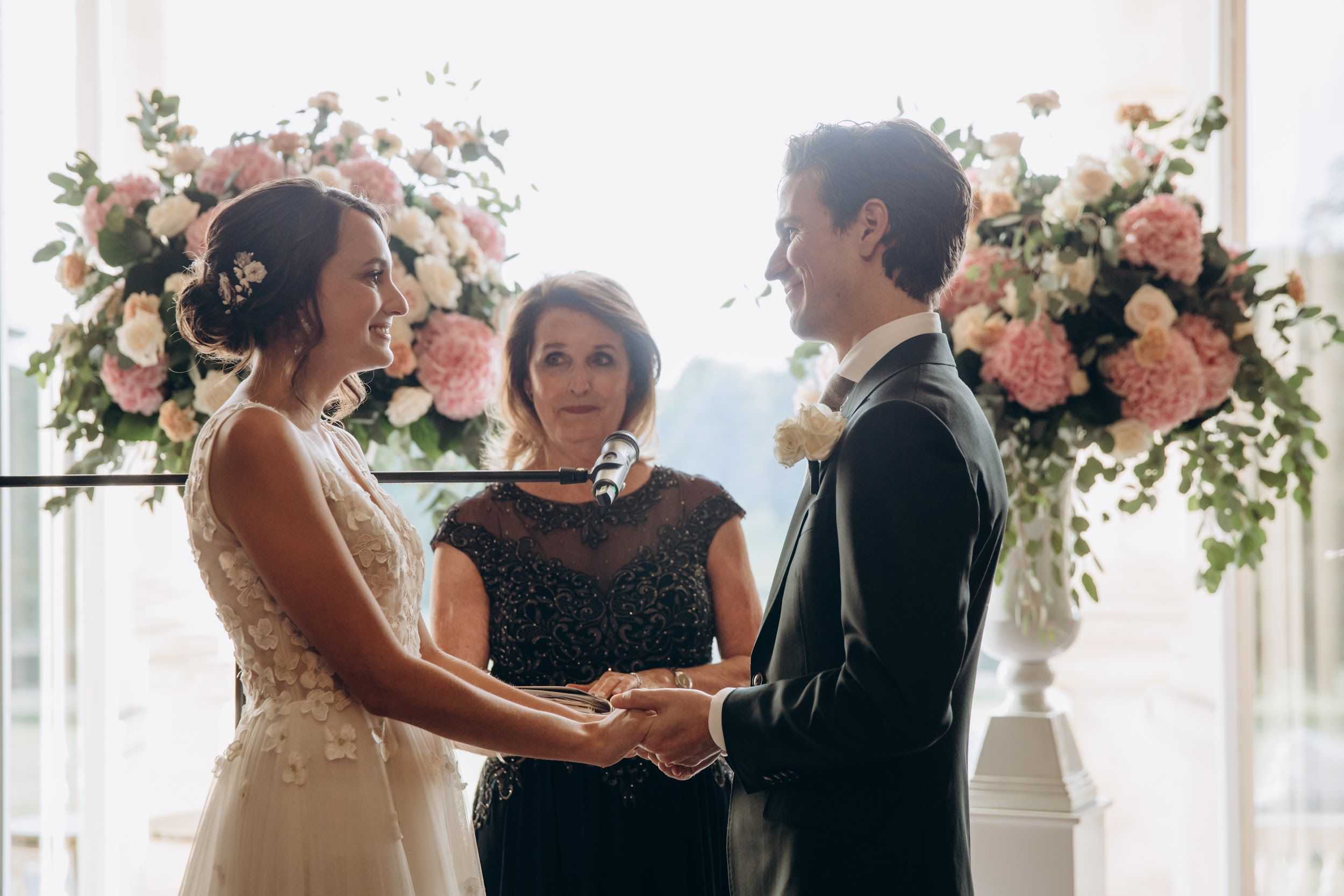 The bride and groom exchange vows during an indoor ceremony, holding hands and smiling at each other while an officiant stands between them at a microphone. The bride wears an ivory gown with three-dimensional floral appliqué detailing and has her dark hair pulled up in a low chignon with small floral hair pins. The groom wears a dark navy suit with a white shirt and an ivory floral boutonnière. The officiant is dressed in a dark beaded cap-sleeve top. Two large floral arrangements in white urn-shaped pedestals frame the altar, composed of blush pink peonies, ivory roses, peach garden roses, and trailing greenery. Large floor-to-ceiling windows behind the altar flood the scene with natural backlight. The shot is a medium portrait framing all three figures from roughly the waist up.