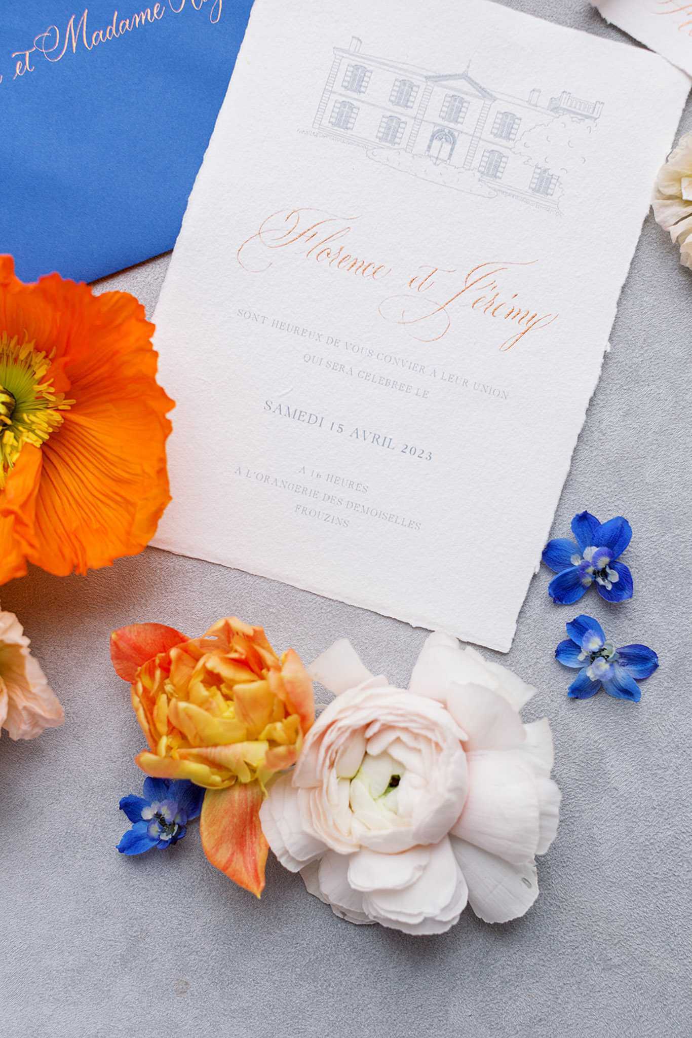A close-up flat lay of a French wedding invitation suite for Florence and Jérémy, dated Saturday April 15, 2023, at L'Orangerie des Demoiselles in Frouzins. The invitation is printed on textured deckle-edge white paper with copper/rose gold calligraphy script for the couple's names and a delicate pencil-style illustrated chateau at the top, paired with a cobalt blue envelope featuring rose gold script addressing. Scattered around the stationery are an orange poppy, an orange and yellow parrot tulip, blush ranunculus, white garden roses, and loose cobalt blue delphinium florets, creating a vibrant color palette of orange, cobalt blue, and blush against a light grey surface.