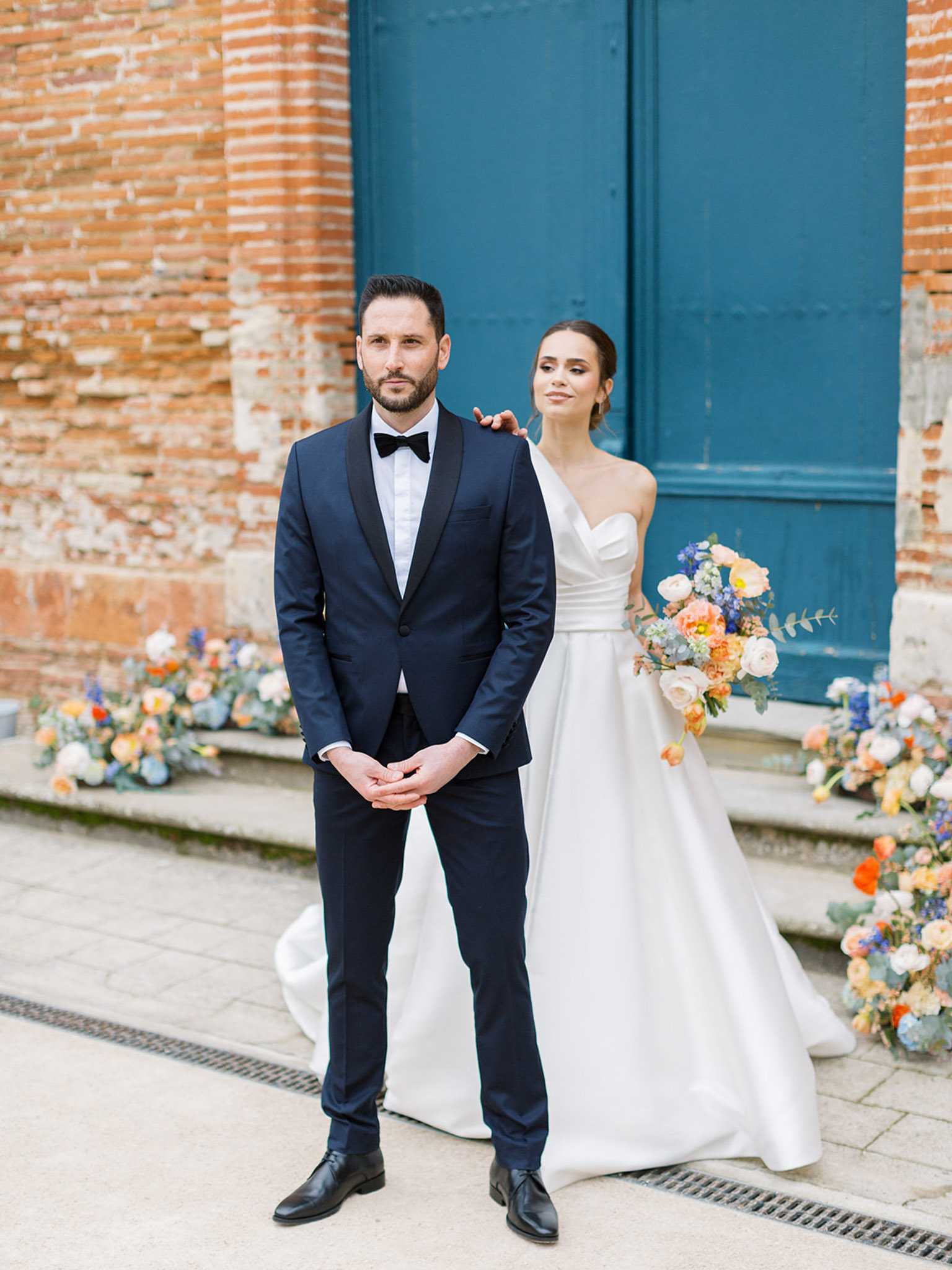 A couple portrait taken outdoors in front of a large teal-painted double door set into an exposed terracotta brick facade. The groom stands slightly in front wearing a navy tuxedo with black satin lapels, a white dress shirt, and a black bow tie, with his hands clasped in front. The bride stands behind him with one hand resting on his shoulder, wearing a strapless ivory ball gown with a structured sweetheart neckline and full satin skirt. She holds a loosely arranged bouquet featuring peach garden roses, coral ranunculus, cobalt blue delphinium, ivory blooms, and eucalyptus. Low floral arrangements in the same color palette — peach, orange, cobalt blue, and ivory — are placed on the steps flanking the couple on both sides, creating a coordinated ground-level floral installation. The overall styling is modern and classic with a bold, warm-toned floral palette. Medium portrait shot.
