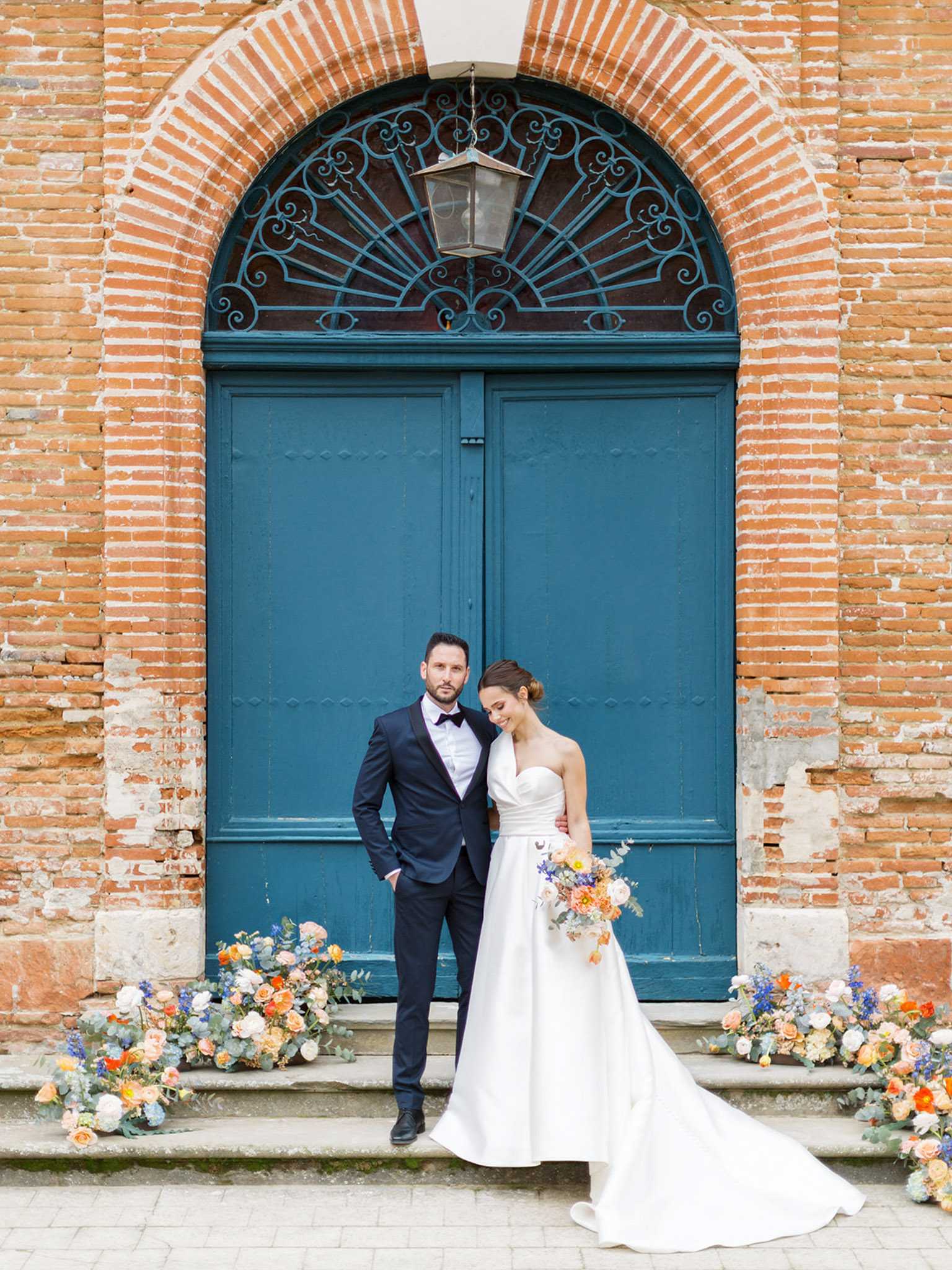 A couple portrait taken outdoors in front of a large teal-painted double door set within a rounded brick archway, with decorative wrought-iron fanlight detailing and a hanging lantern above. The groom wears a navy tuxedo with a black bow tie, and the bride wears a strapless white ball gown with a structured bodice and a full skirt with a trailing hem, her hair pulled back in a low bun. She holds a loose, garden-style bouquet of peach and orange roses, blue delphinium, white ranunculus, and eucalyptus foliage, and looks downward while the groom faces the camera with his arm around her. Clustered floral arrangements in matching peach, orange, blue, and white tones with eucalyptus are placed along the base of the doorstep on both sides, reinforcing a warm, earthy color palette with cool blue accents. The composition is a full-length portrait shot with the architectural doorway serving as a strong graphic backdrop.