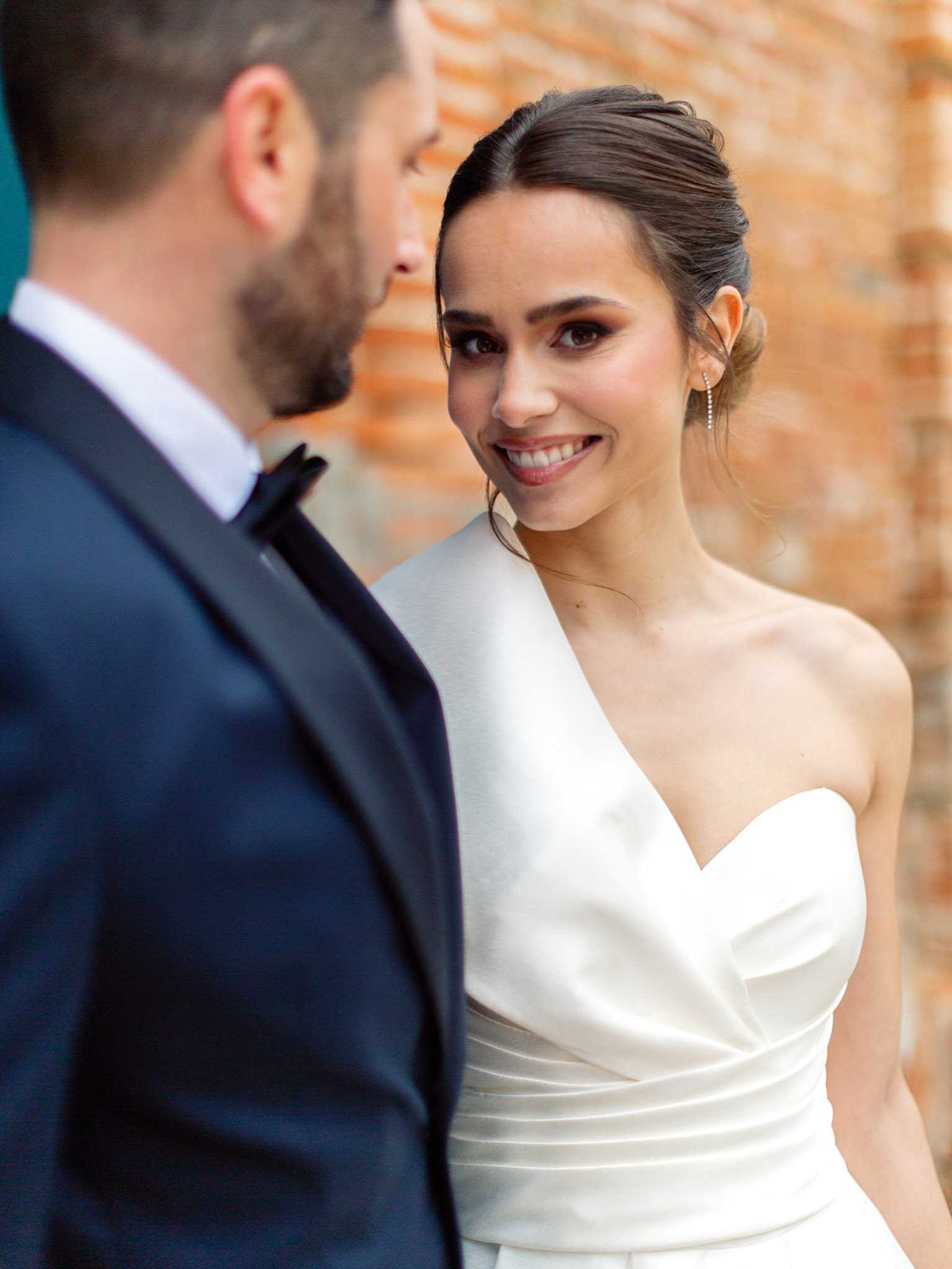 A couple portrait shot outdoors, with the bride in sharp focus smiling at the groom, who is blurred in the foreground. The bride wears a white satin one-shoulder structured gown with pleated bodice detailing and a sweetheart neckline, her dark hair styled in a low updo with delicate drop earrings. The groom is dressed in a navy suit with a black bow tie. The background features a brick wall, softly out of focus. The styling is clean and modern with a minimalist dress silhouette. Portrait composition with shallow depth of field.