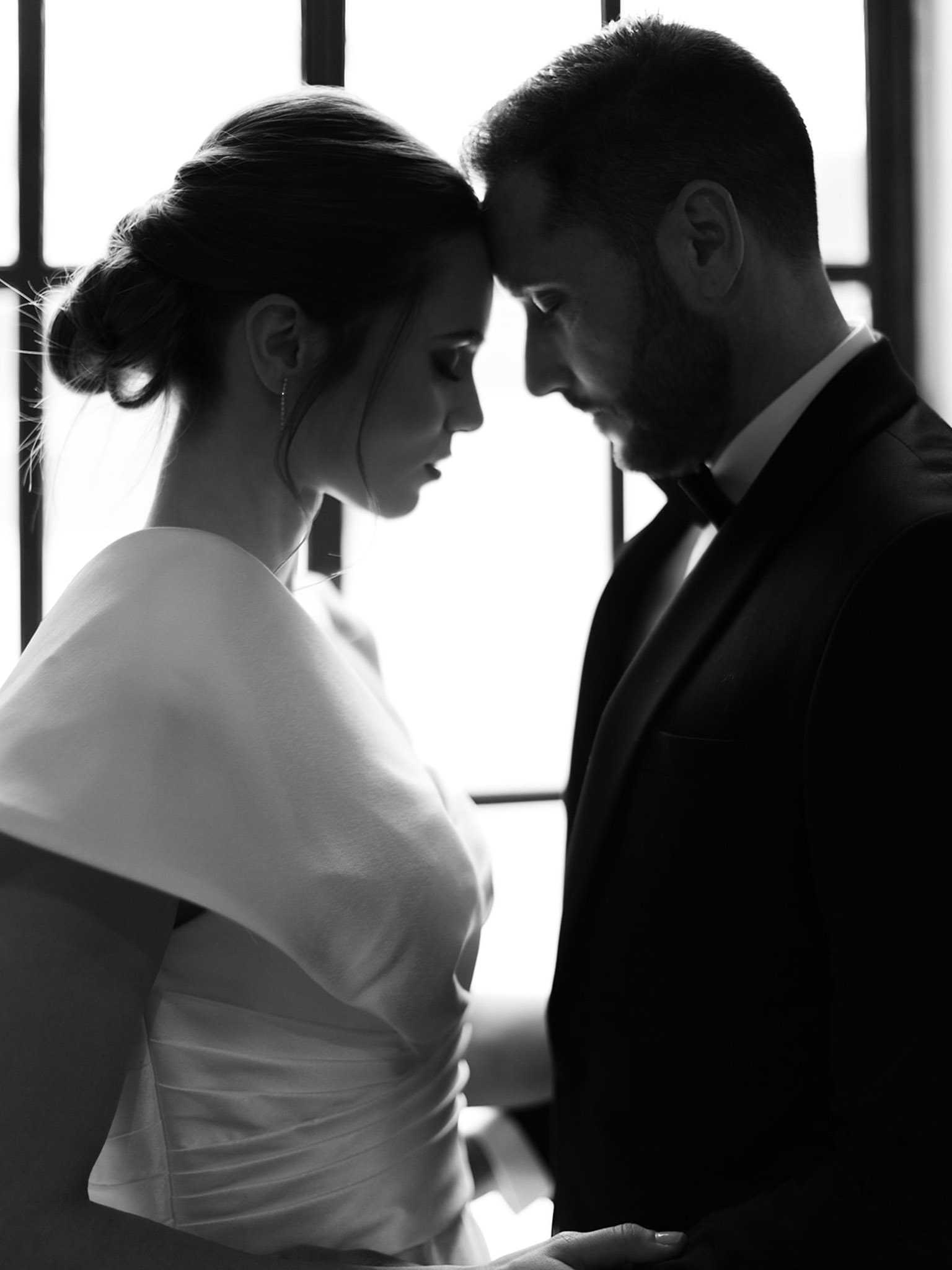 A black-and-white couple portrait shot in profile, with the bride and groom standing face to face with their foreheads nearly touching. The bride wears a minimalist white gown with a structured off-the-shoulder or cape-style bodice and has her hair styled in a low updo, with a delicate drop earring visible. The groom wears a dark tuxedo with a white dress shirt and black bow tie, and has a short beard. The image is backlit by large window frames behind them, creating strong contrast between the bright white background and the deeply shadowed figures. The composition is a tight upper-body portrait emphasizing the couple's closeness and the clean lines of their attire.