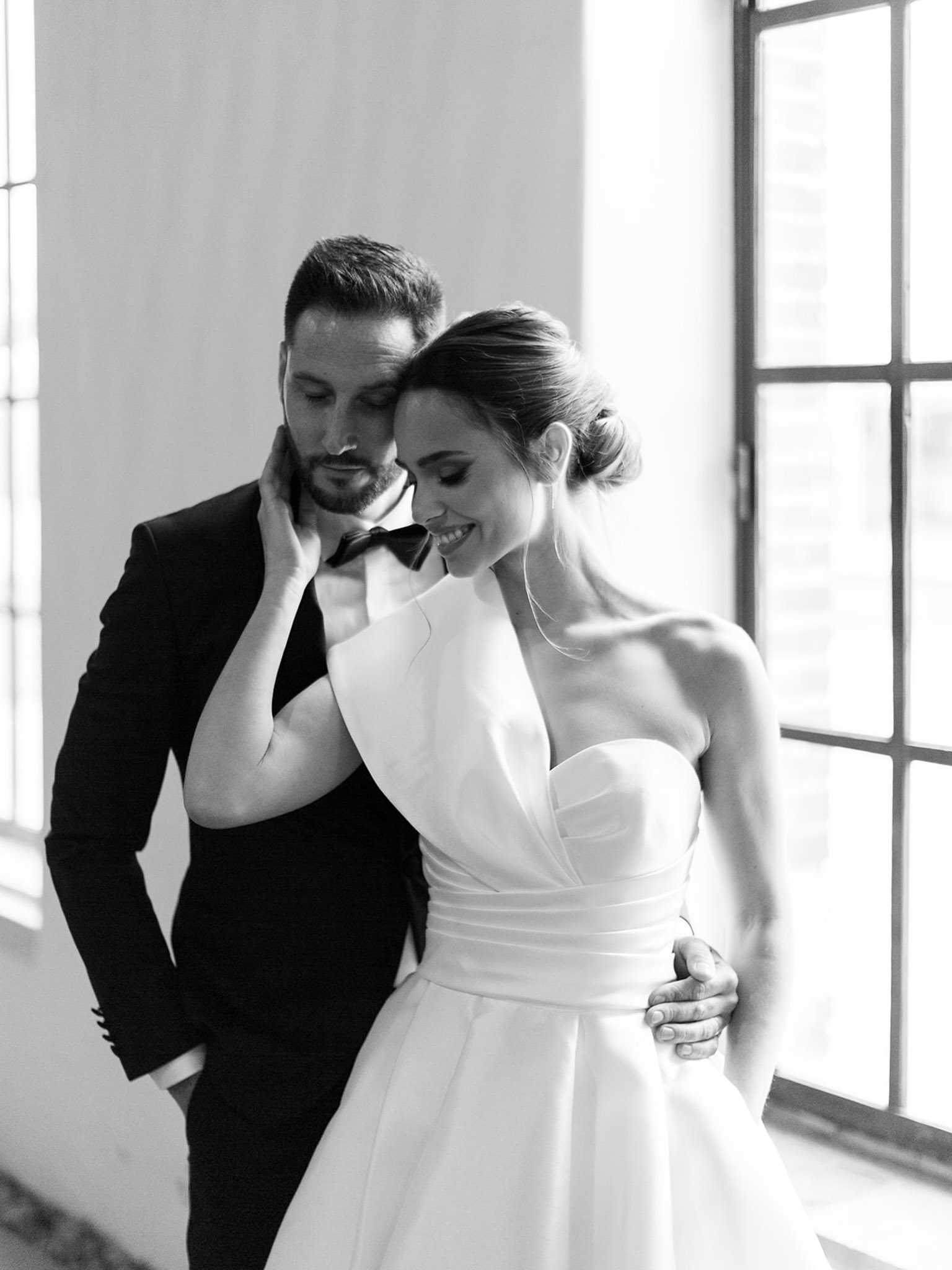 This is a black-and-white couple portrait taken indoors, with soft natural light streaming through large steel-framed windows in the background. The bride wears a structured, architectural strapless gown with a sculptural asymmetric folded bodice and a defined waistband, styled with a low chignon updo; the groom is dressed in a dark tuxedo with a bow tie. The couple stands close together, the groom positioned slightly behind with his arm around the bride's waist, while she smiles downward and rests her hand gently against his jaw. The image has bright highlights and soft mid-tones with a clean, modern styling aesthetic, shot as a close-cropped portrait with shallow depth of field.