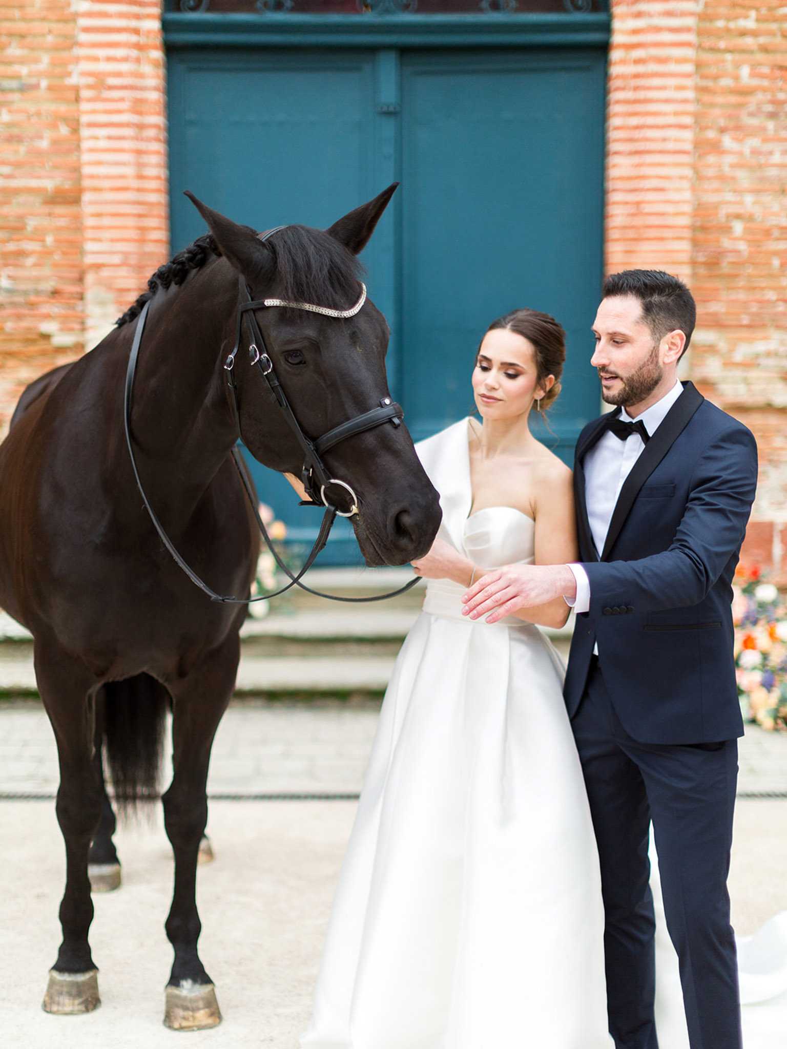 A couple portrait taken outdoors in the courtyard of a red brick building featuring a large teal double door. The bride wears a strapless white ball gown with a structured bodice and a white wrap or stole over her shoulders, with her hair in an updo, while the groom wears a navy tuxedo with black lapels and a black bow tie. Both are petting a dark bay horse fitted with a black leather bridle adorned with a rhinestone browband and a braided mane. A colorful floral arrangement is partially visible in the background to the right. The setting has a classic, formal aesthetic. Medium portrait shot.