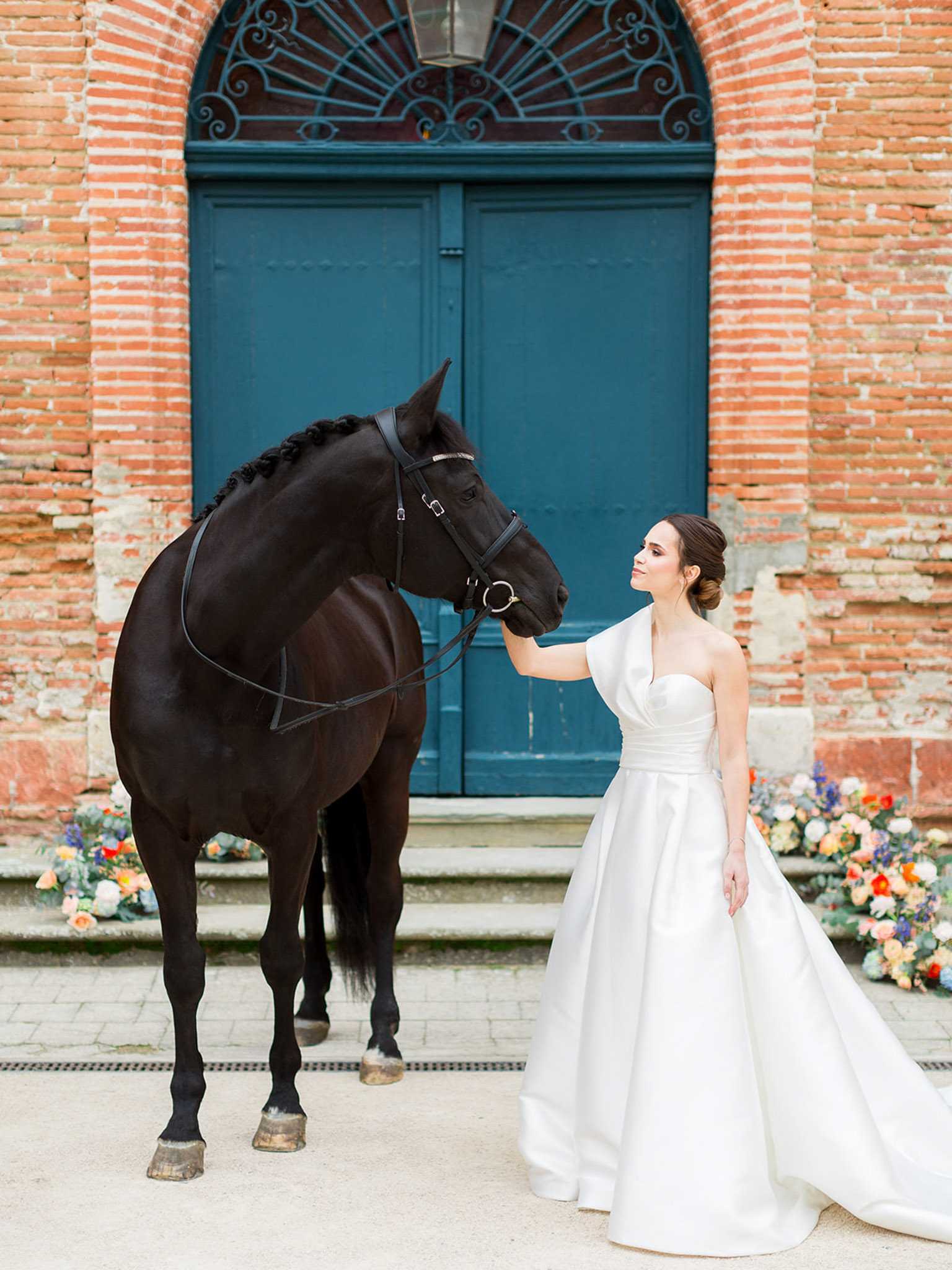 A bridal portrait taken outdoors in front of a grand teal double door set within a red brick arched entrance, likely at a French chateau or estate. The bride wears a white structured ballgown with a one-shoulder draped detail and a full satin skirt, her dark hair styled in a low updo, and she gently holds the bridle of a black horse with a braided mane standing beside her. Low floral arrangements in peach, orange, ivory, and blue-purple tones are placed on the steps flanking the doorway, adding a colorful accent to the classic architectural backdrop. The composition is a full-length portrait with a modern, polished aesthetic.