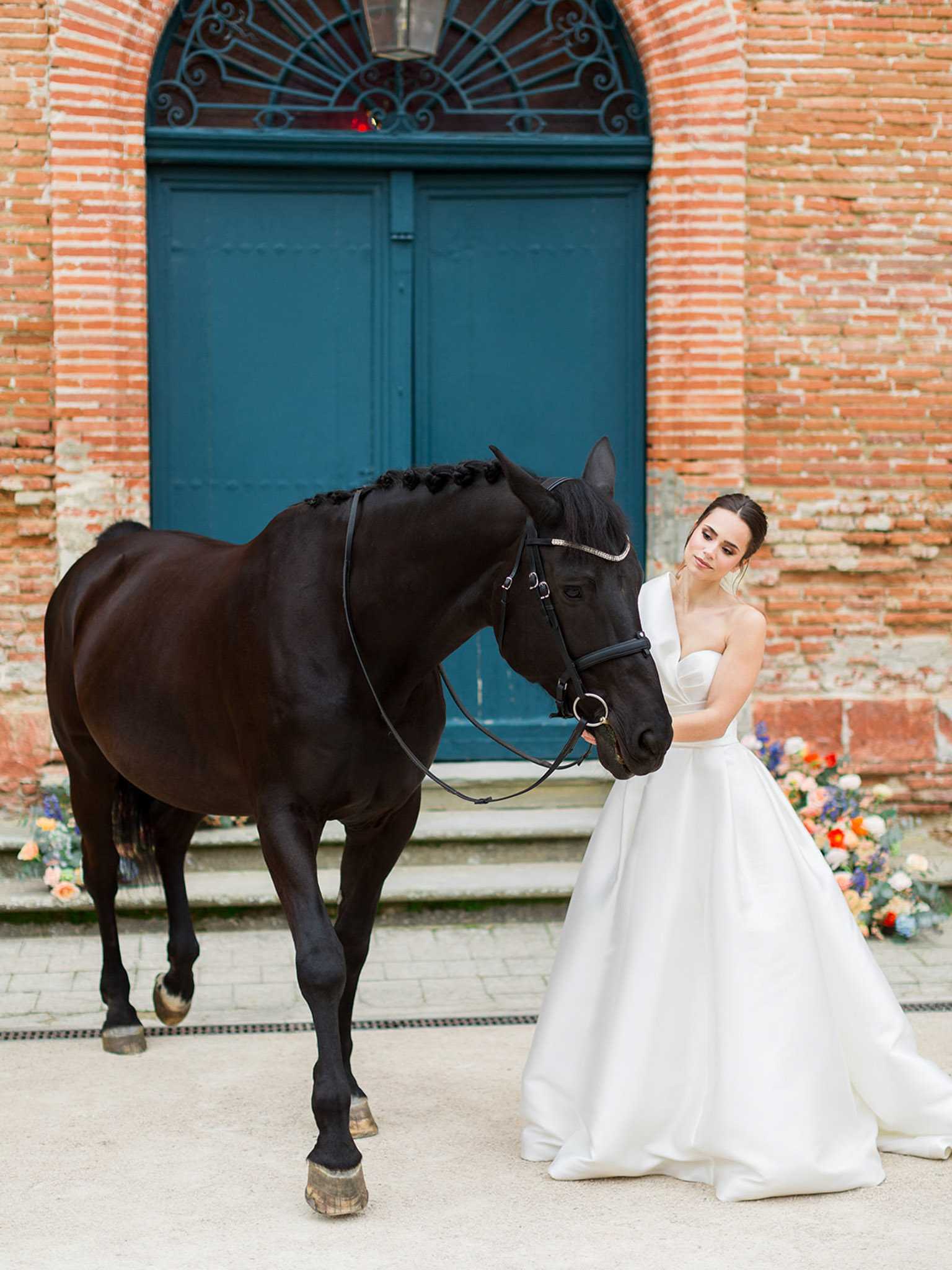 A bridal portrait shot outdoors in front of a red brick building featuring a large teal arched double door with decorative ironwork. The bride wears a white one-shoulder ballgown with a full satin skirt and holds the bridle of a dark brown-black horse whose mane is braided with small dark flowers. She is looking down at the horse's nose, which she is cradling gently with her other hand. Behind her to the right, a loose floral arrangement on the steps features blooms in coral, peach, white, blue, and orange tones. The overall styling is modern and clean. Medium full-length portrait composition.