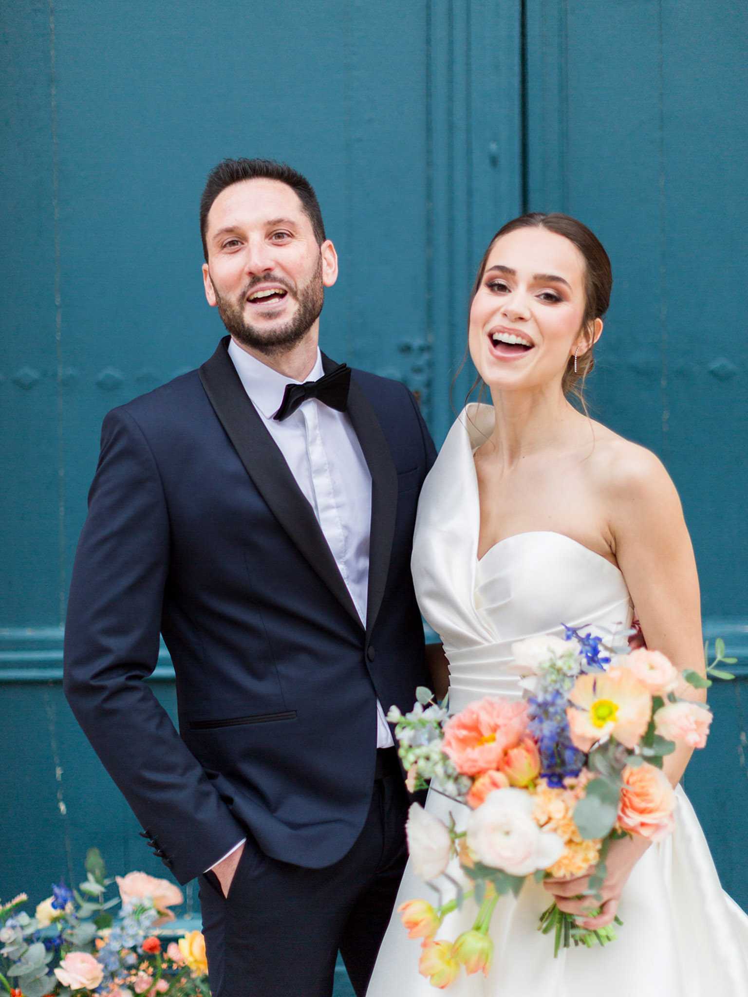 A couple portrait shot outdoors in front of large teal-painted double doors, both laughing openly. The groom wears a navy tuxedo with black satin lapels and a black bow tie. The bride wears a white strapless structured satin gown with a draped asymmetrical neckline detail, her hair pulled back in an updo. She holds a loose, garden-style bridal bouquet featuring peach ranunculus, coral poppies, cobalt blue delphinium, white blooms, yellow accents, and eucalyptus. A matching floral arrangement is visible at ground level in the lower foreground. The overall color palette of the florals is vibrant — peach, coral, cobalt blue, and yellow — against the deep teal door background. Medium portrait crop, shot at chest height.