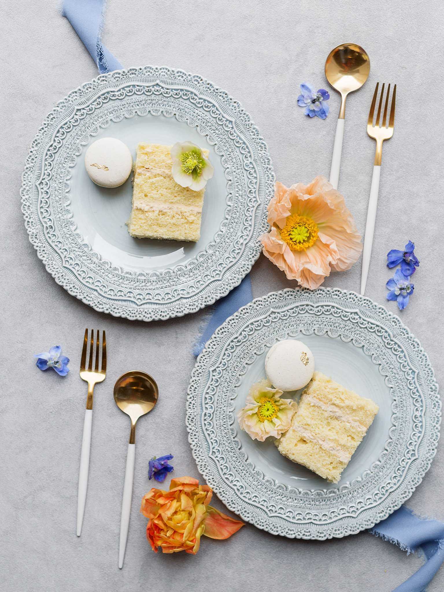 A styled flat-lay detail shot of wedding dessert tableware arranged on a light grey surface. Two pale blue lace-edged ceramic plates each hold a slice of layered white cake, a white macaron with a small gold leaf detail, and a small yellow-centered flower used as garnish. Gold and white two-tone cutlery — a fork and spoon — is placed beside each plate. Scattered across the surface are peach poppies, small blue delphinium florets, and an orange garden rose, along with draped lengths of frayed pale blue silk ribbon. The overall decor palette combines pale blue, white, gold, and soft peach in a refined yet romantic styling.