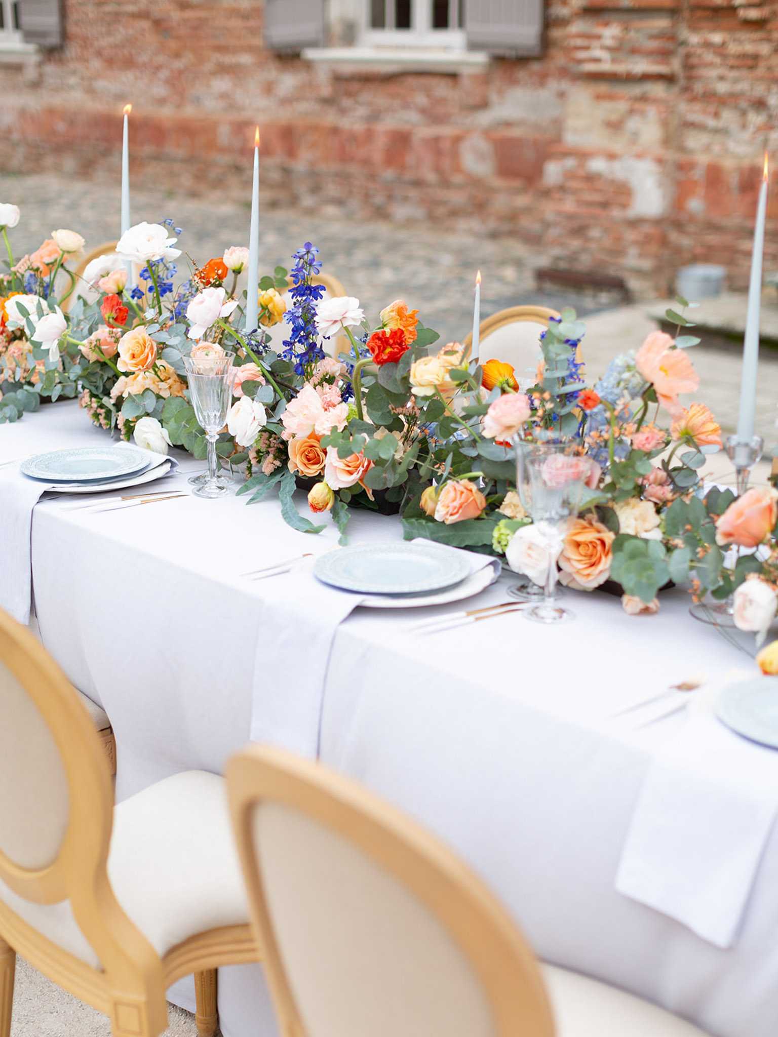 A detail shot of an outdoor wedding reception table set against the brick facade of what appears to be a French chateau or manor. The table is dressed in a pale grey-blue linen with matching pale blue charger plates, crystal glassware including fluted champagne glasses, and rose gold flatware. A lush floral runner runs the full length of the table, composed of peach and apricot garden roses, white ranunculus, orange spray roses, cobalt blue delphiniums, dusty blue scabiosa, and trailing eucalyptus. Pale blue taper candles are lit and interspersed throughout the floral arrangement. The chairs are natural wood Louis XVI-style with cream upholstered seats. The overall decor palette combines warm peach and orange tones with cool dusty blue accents in a classic French style.