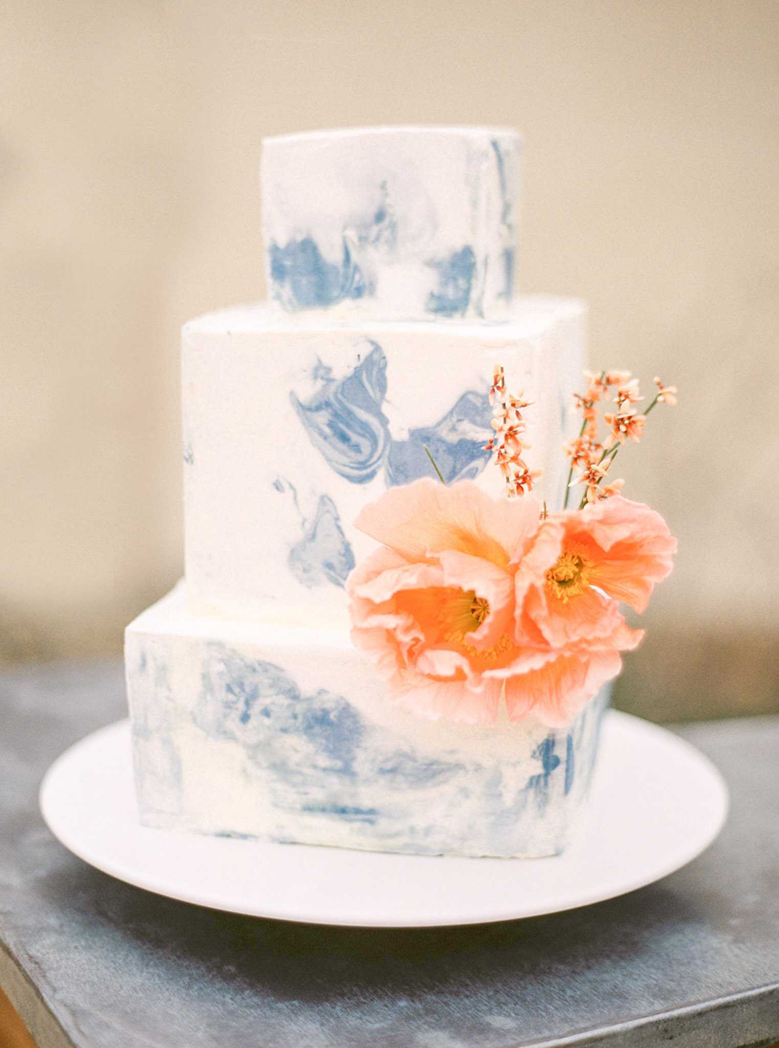 Close-up detail shot of a three-tier wedding cake displayed on a white round cake board atop a grey surface. The cake features a white buttercream base with hand-painted indigo blue marbling across all three tiers, which alternate between a square bottom tier, a round middle tier, and a smaller round top tier. The cake is decorated with two large coral-peach poppy blooms with yellow centers and small sprigs of delicate coral wildflowers. The overall design palette combines indigo blue marbling with coral and peach florals against a neutral cream background.