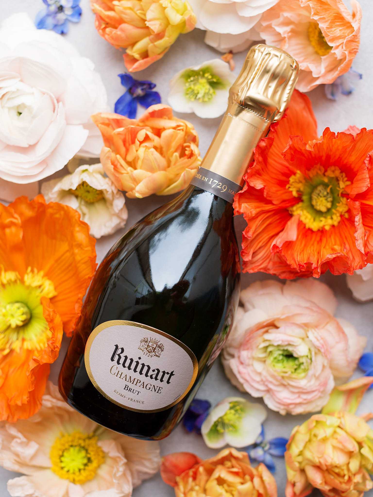 A flat lay detail shot featuring a bottle of Ruinart Brut Champagne (Reims, France) laid diagonally on a light surface surrounded by an arrangement of loose flower heads. The floral palette includes orange and coral Icelandic poppies, peach and apricot parrot tulips, blush and white ranunculus, small blue delphinium florets, and white hellebores. The bottle has a dark green glass body, gold foil capsule, and a cream label with gold border. No people are present; the image is styled as a product or wedding detail shot with a vibrant, garden-inspired color palette.