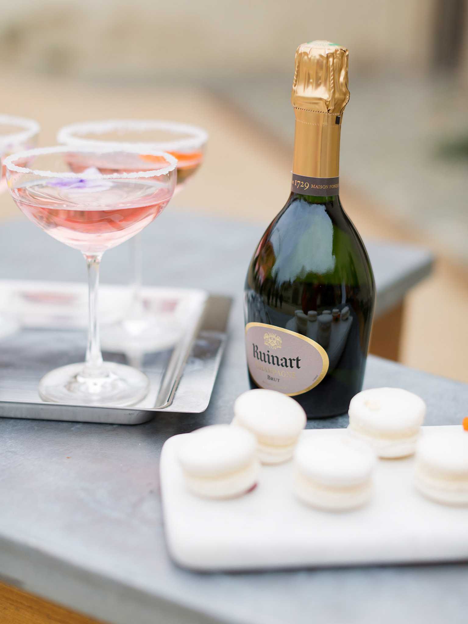 A close-up detail shot of a cocktail hour beverage and dessert display set on a grey stone surface. A bottle of Ruinart Brut champagne with its gold foil and distinctive label is positioned centrally, flanked by coupe glasses filled with a pink rosé-tinted cocktail and rimmed with sugar, resting on a silver tray alongside a cocktail stirrer. In the foreground, a white marble board holds six ivory-white French macarons arranged in two rows. The background is softly blurred, suggesting an outdoor or semi-outdoor setting. The overall styling is classic and refined, with a pale neutral palette of ivory, blush, and grey.