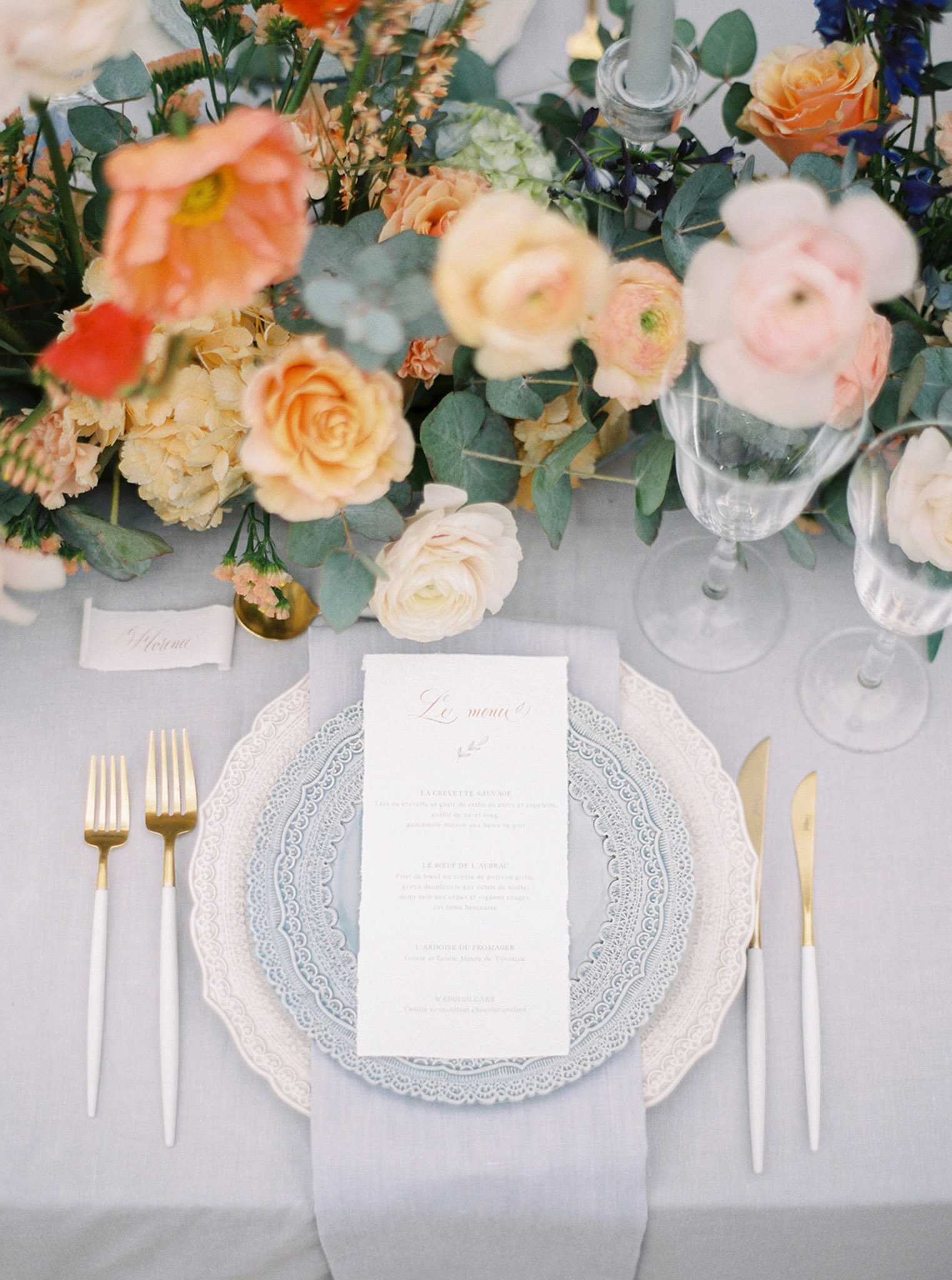 A close-up flat lay detail shot of a wedding reception place setting on a dusty blue-grey linen tablecloth. The place setting features a pale blue decorative charger plate with a white lace-edged rim, topped with a white deckle-edge French menu card reading 'Le menu' in coral-red calligraphy script, flanked by white and gold flatware with two forks on the left and a knife on the right. Two clear glass goblets sit to the upper right alongside a small white place card with calligraphed script. The upper portion of the image is filled with a lush floral centerpiece composed of coral poppies, peach and apricot garden roses, cream hydrangeas, blush and pink ranunculus, cobalt blue delphinium, dusty miller, eucalyptus, and small peach wax flowers, all arranged in a loose, organic garden-style design with grey-blue taper candles visible in the background.