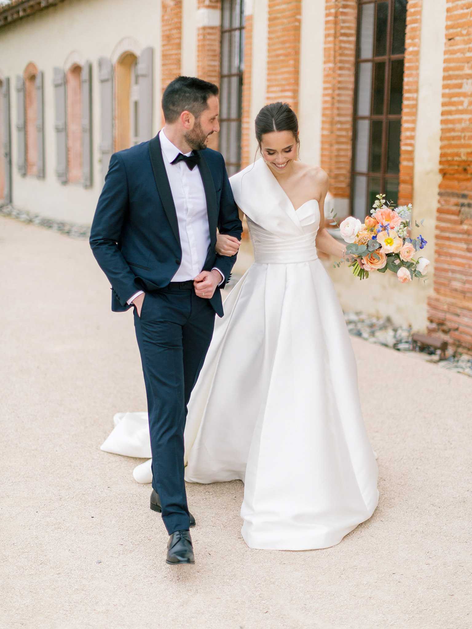A couple portrait taken outdoors on a gravel path alongside a red brick building with grey shuttered windows, consistent with a French chateau or domaine. The bride wears a structured ivory satin ballgown with a one-shoulder draped neckline and a short train, and carries a loose bouquet of peach garden roses, coral tulips, white ranunculus, blue delphinium, and eucalyptus foliage. The groom wears a navy tuxedo with black satin lapels, a white dress shirt, and a black bow tie. The two are walking arm in arm, the groom looking toward the bride while she smiles downward, in a candid full-length portrait composition.