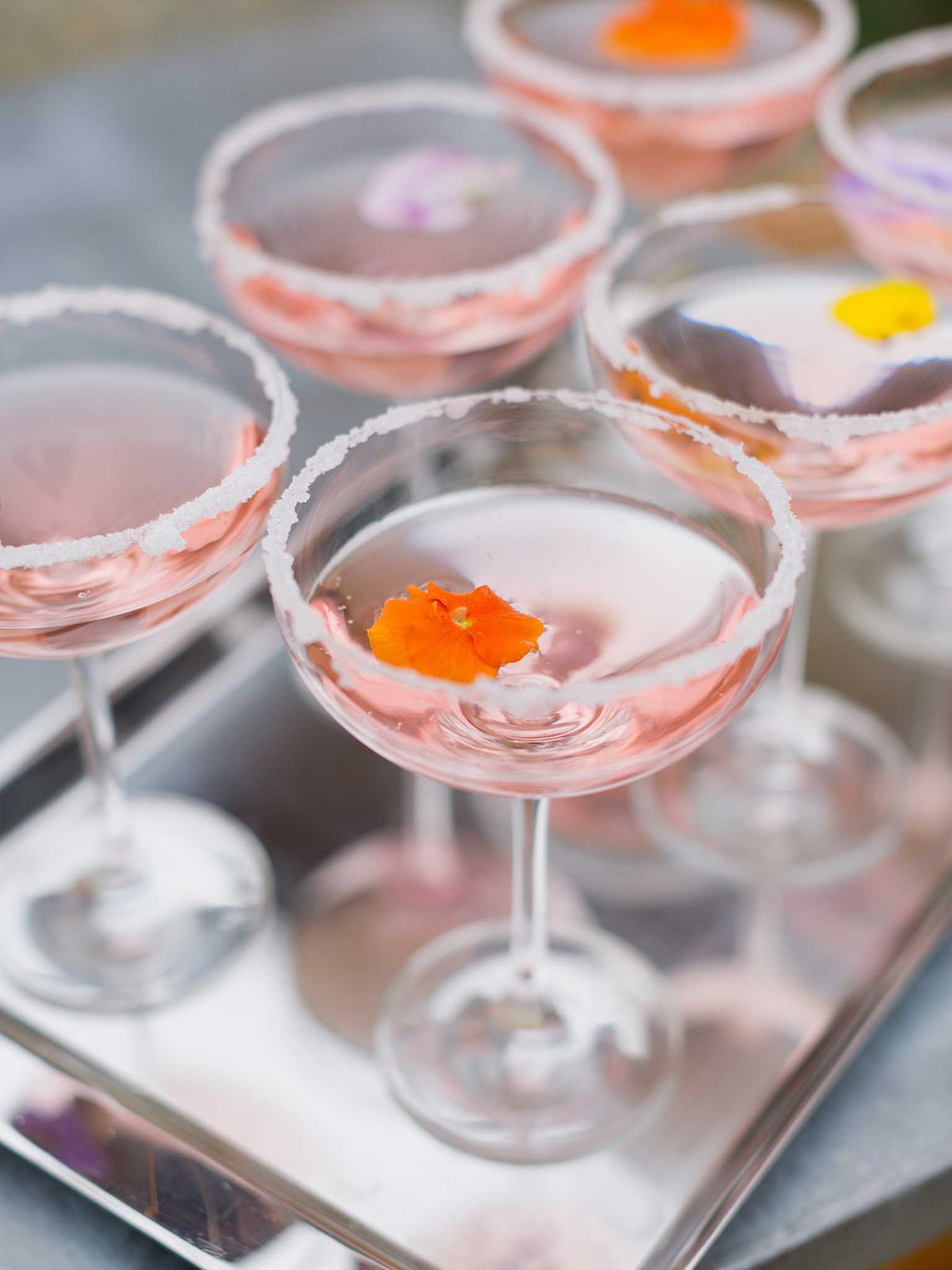 Close-up detail shot of cocktail hour drinks displayed on a silver rectangular tray. Five coupe-style glasses are filled with a pale blush pink cocktail, each rimmed with white sugar and garnished with a single edible flower floating on the surface — including an orange nasturtium, a yellow flower, and small purple blooms. The styling is refined and intentional, with the floral garnishes adding pops of orange, yellow, and lavender against the soft pink drinks. The composition uses a shallow depth of field, with the foreground glass in sharp focus and the remaining glasses softly blurred.