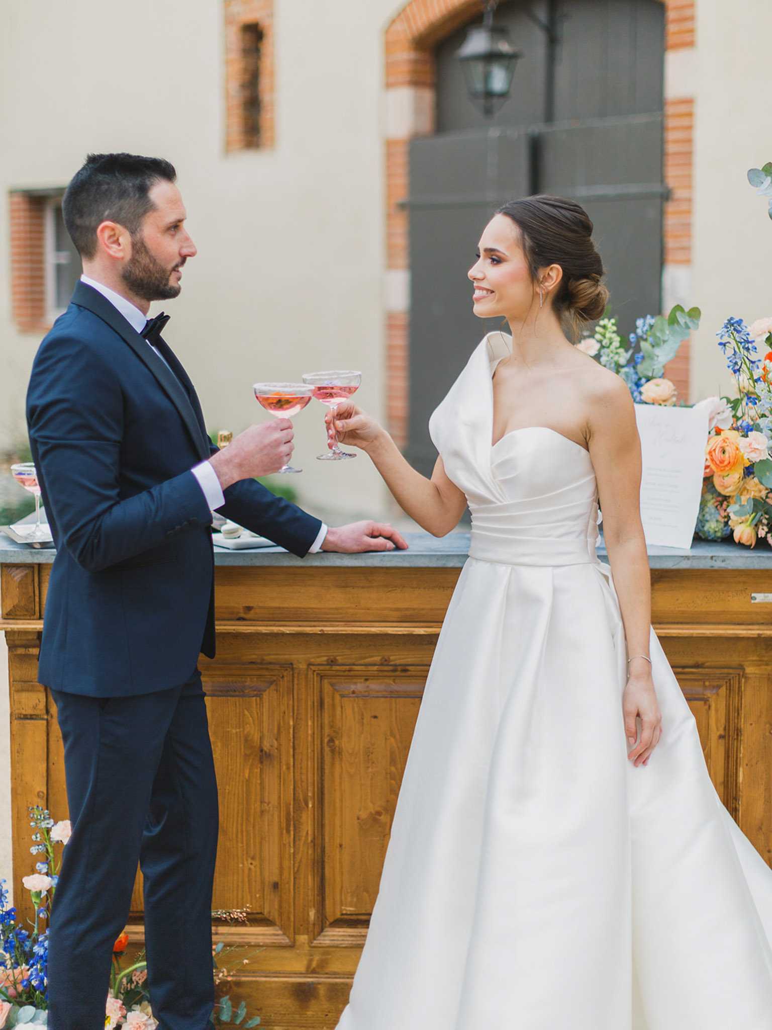 A couple toasts with pink coupe glasses at an outdoor cocktail hour, standing beside a rustic wooden bar with a zinc or slate top. The groom wears a fitted navy suit with a black bow tie, while the bride wears a structured ivory satin ball gown with a one-shoulder ruched bodice and full skirt, her hair styled in an updo. Floral arrangements in cobalt blue delphiniums, peach and orange ranunculus, and eucalyptus are visible on and around the bar, suggesting a color palette of blue, peach, and terracotta. The setting appears to be a French chateau or domaine courtyard, with rendered walls and red brick detailing visible in the background; the shot is a mid-length portrait of the couple facing each other.