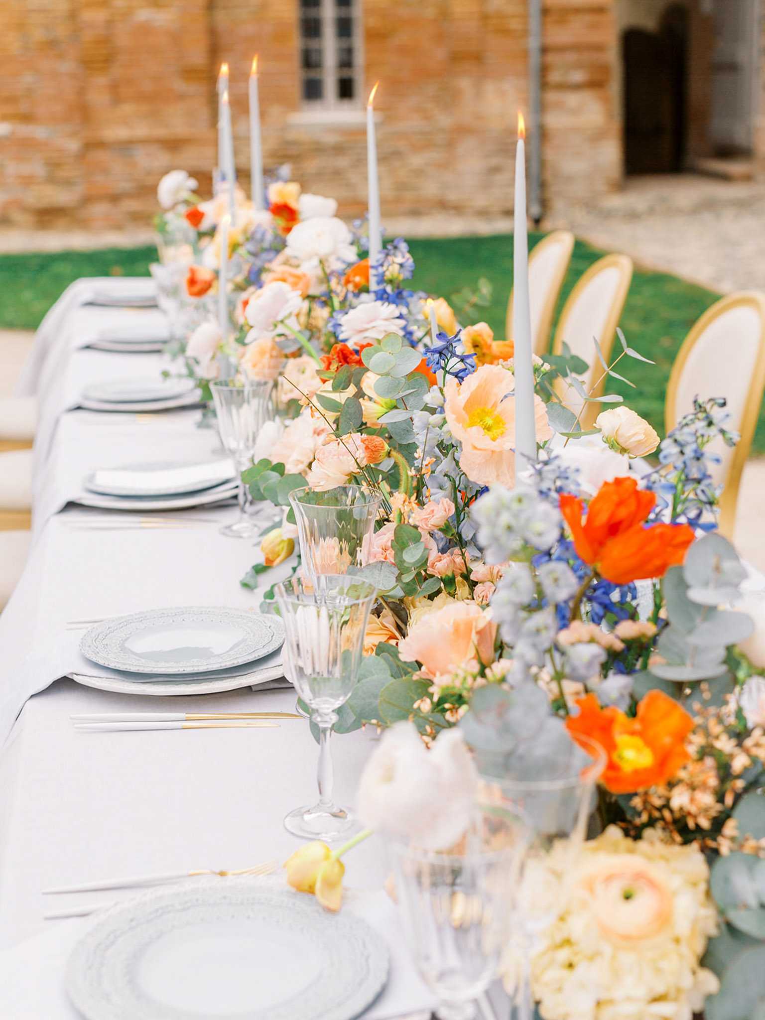 A close-up detail shot of an outdoor wedding reception table set against the stone façade of what appears to be a French chateau or manor. The long rectangular table is dressed in a pale grey linen with place settings of layered grey charger plates and white dinner plates, gold-toned flatware, and crystal glassware. A lush floral runner runs the length of the table, composed of orange Iceland poppies, peach garden roses, blush ranunculus, blue delphinium, dusty blue scabiosa, and eucalyptus foliage. Slim grey taper candles in varying heights are lit and interspersed throughout the floral arrangement, adding vertical structure. Gold bamboo-style chairs are visible along one side of the table. The overall decor palette is grey, peach, orange, and blue, with a classic French garden aesthetic.