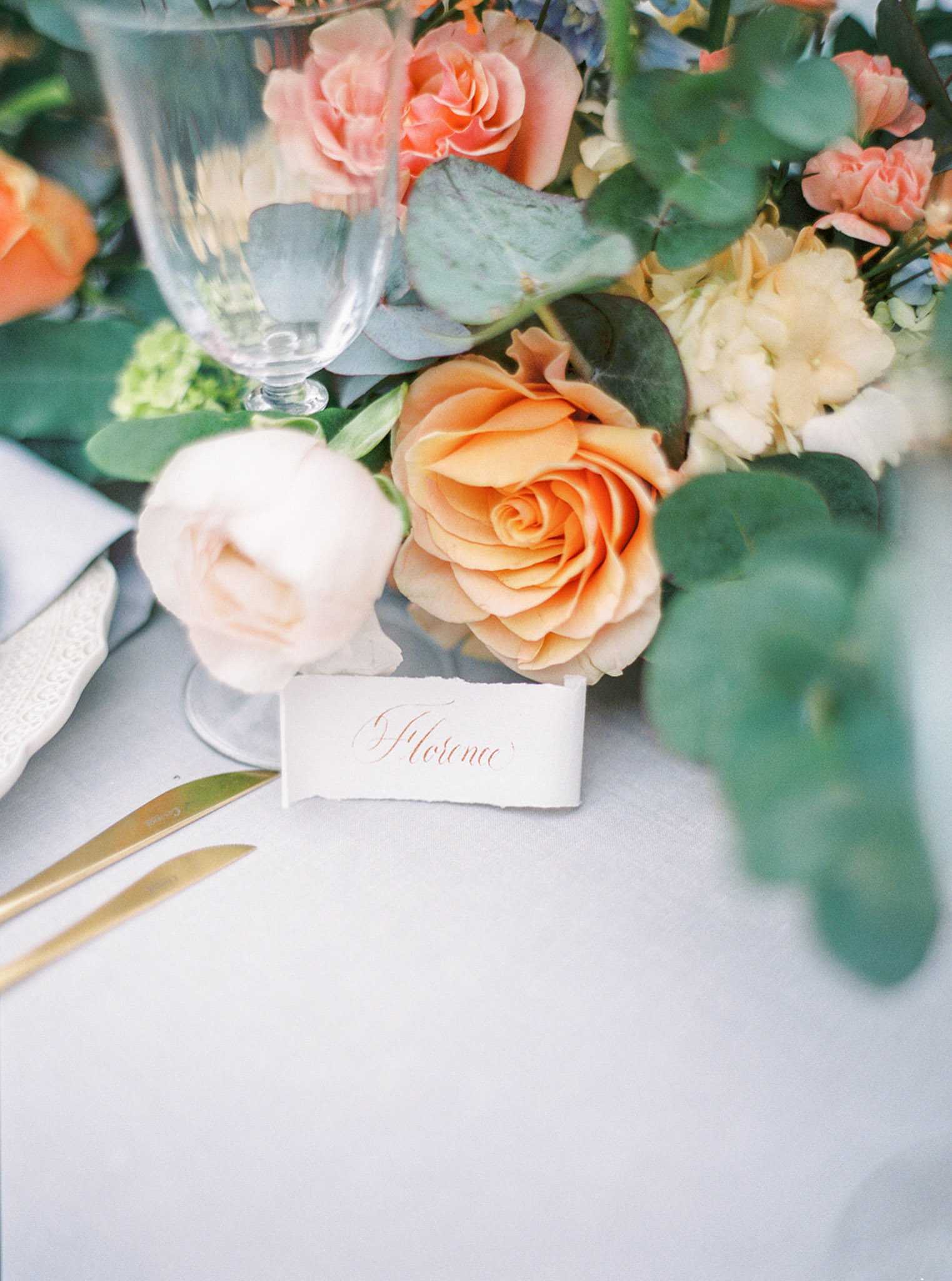 Close-up detail shot of a wedding reception place setting featuring a hand-torn deckle-edge place card with the name 'Florence' written in copper-toned calligraphy script. Gold flatware is laid on a light grey linen tablecloth alongside a clear crystal wine glass and a decorative silver-patterned knife. The floral centerpiece spills into the frame with peach and coral garden roses, ivory hydrangea, soft white ranunculus, touches of blue flowers, and eucalyptus foliage, creating a warm peach, ivory, and green palette with a classic romantic styling.
