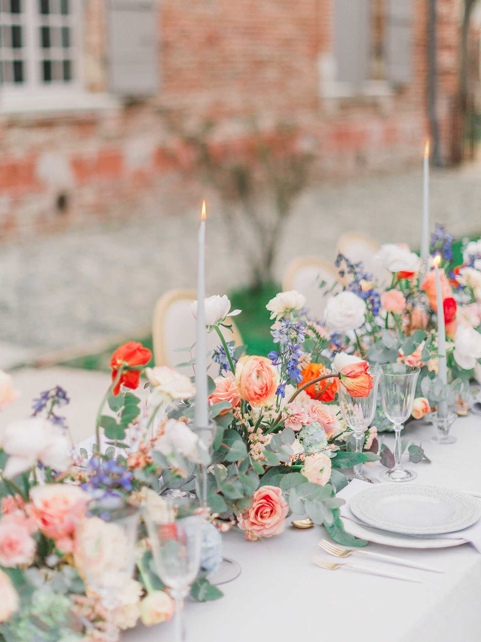 A close-up detail shot of an outdoor reception table set against the blurred façade of a brick building, likely a French chateau or manor. The white linen table is dressed with a lush floral runner composed of coral and peach ranunculus, red poppies, ivory garden roses, blush roses, periwinkle blue delphinium, and eucalyptus foliage. Tall, pale grey-blue taper candles are lit and placed at intervals throughout the florals, and place settings feature white embossed charger plates stacked with a smaller white plate, gold flatware, and crystal glassware including a champagne flute and wine glass. The overall decor palette combines soft grey-blue linens and candles with a vibrant, garden-style floral arrangement in warm coral, red, peach, and blush tones accented by blue and white blooms, creating a classic French garden aesthetic.