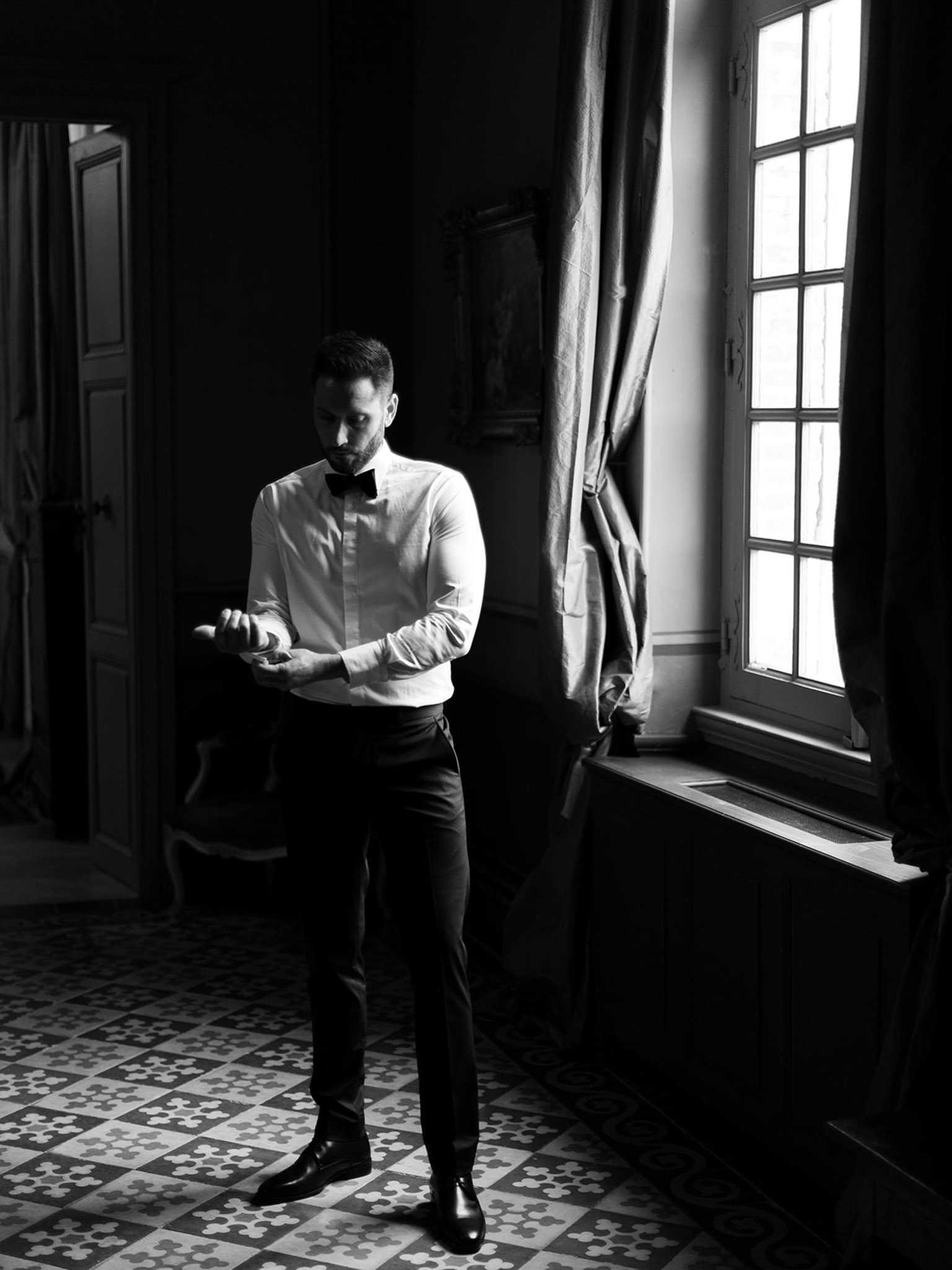 Black-and-white full-length portrait of a groom getting ready indoors, adjusting his cufflinks while standing in a dimly lit room beside a tall multi-pane window. He is dressed in a white dress shirt, black bow tie, dark trousers, and black leather dress shoes. The image is high contrast, with bright natural light streaming in from the window creating deep shadows across the room. The floor features a decorative geometric patterned tile, and heavy draped curtains frame the window, with an open panelled door and a framed painting partially visible in the background, suggesting a classic French chateau interior.