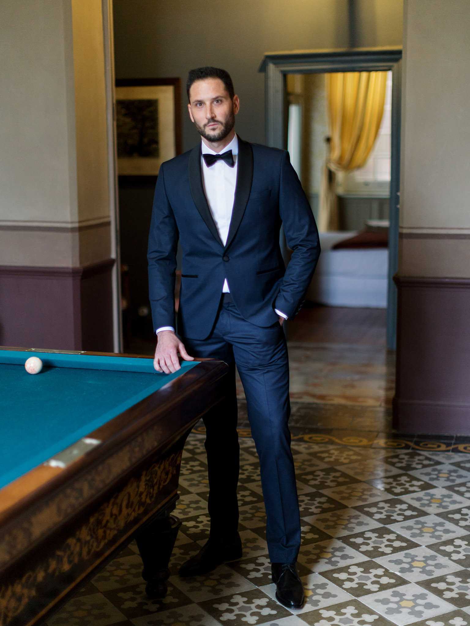 A groom portrait taken indoors in what appears to be a billiards room within a chateau or manor house. The groom stands beside a pool table with teal felt, wearing a navy blue tuxedo with black satin shawl lapels, a white dress shirt, and a black bow tie, with one hand resting on the table and the other in his pocket. The room features ornate patterned encaustic floor tiles in grey, white, and brown, pale walls with a mauve lower panel, and open doorways leading to further rooms — one of which reveals golden yellow curtains. The composition is a full-length portrait shot at eye level.