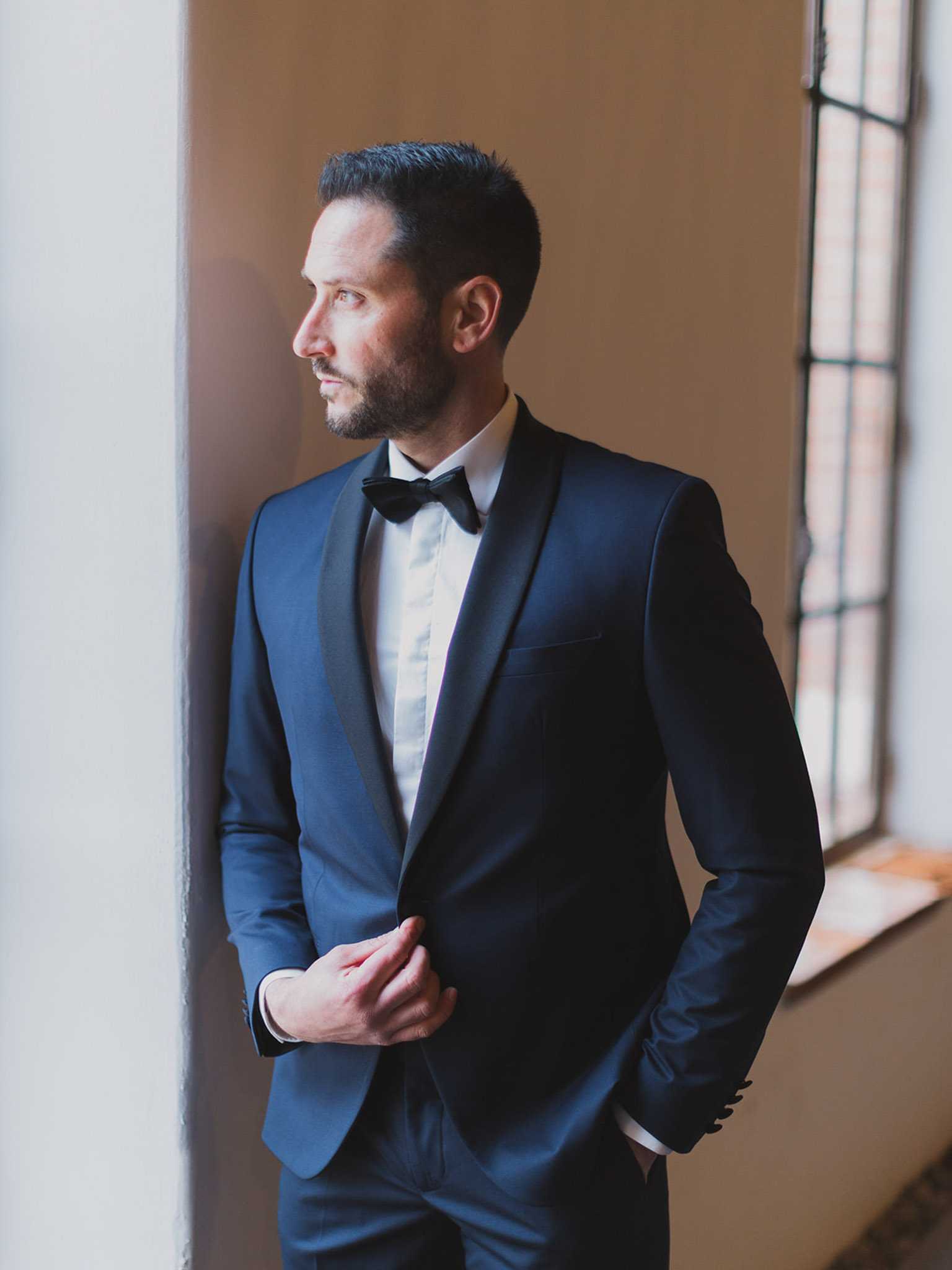 A getting-ready portrait of the groom standing alone indoors beside a tall window with iron-grid detailing, gazing to the side with one hand holding the lapel of his jacket. He is dressed in a navy blue tuxedo with black satin shawl lapels, a white dress shirt, and a black bow tie. The composition is a mid-length portrait framed by soft natural window light on one side and a plain warm-toned wall on the other, creating a classic, formal styling aesthetic.