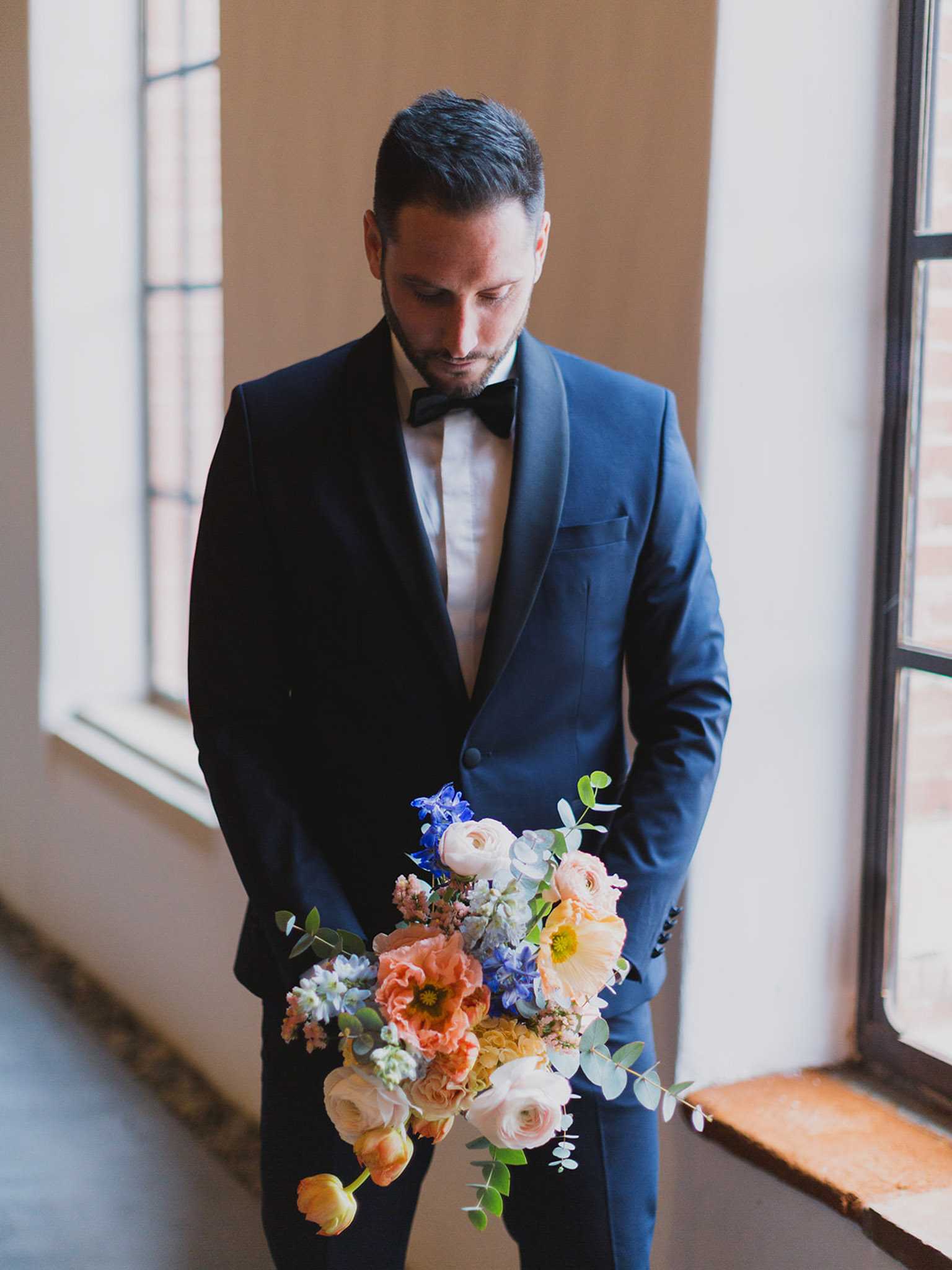 A portrait of the groom standing indoors near large windows, looking down at a bridal bouquet he holds at his side. He is dressed in a fitted navy blue tuxedo with a shawl lapel, white dress shirt, and black bow tie. The bouquet is a loose, garden-style arrangement featuring coral and peach poppies, blush and cream ranunculus, yellow tulips, blue delphinium, white hyacinth, dusty pink stock, and eucalyptus foliage. Natural window light illuminates the scene against a plain plaster wall, giving the image a clean, modern aesthetic. The shot is a medium portrait framed from roughly knee height to above the head.