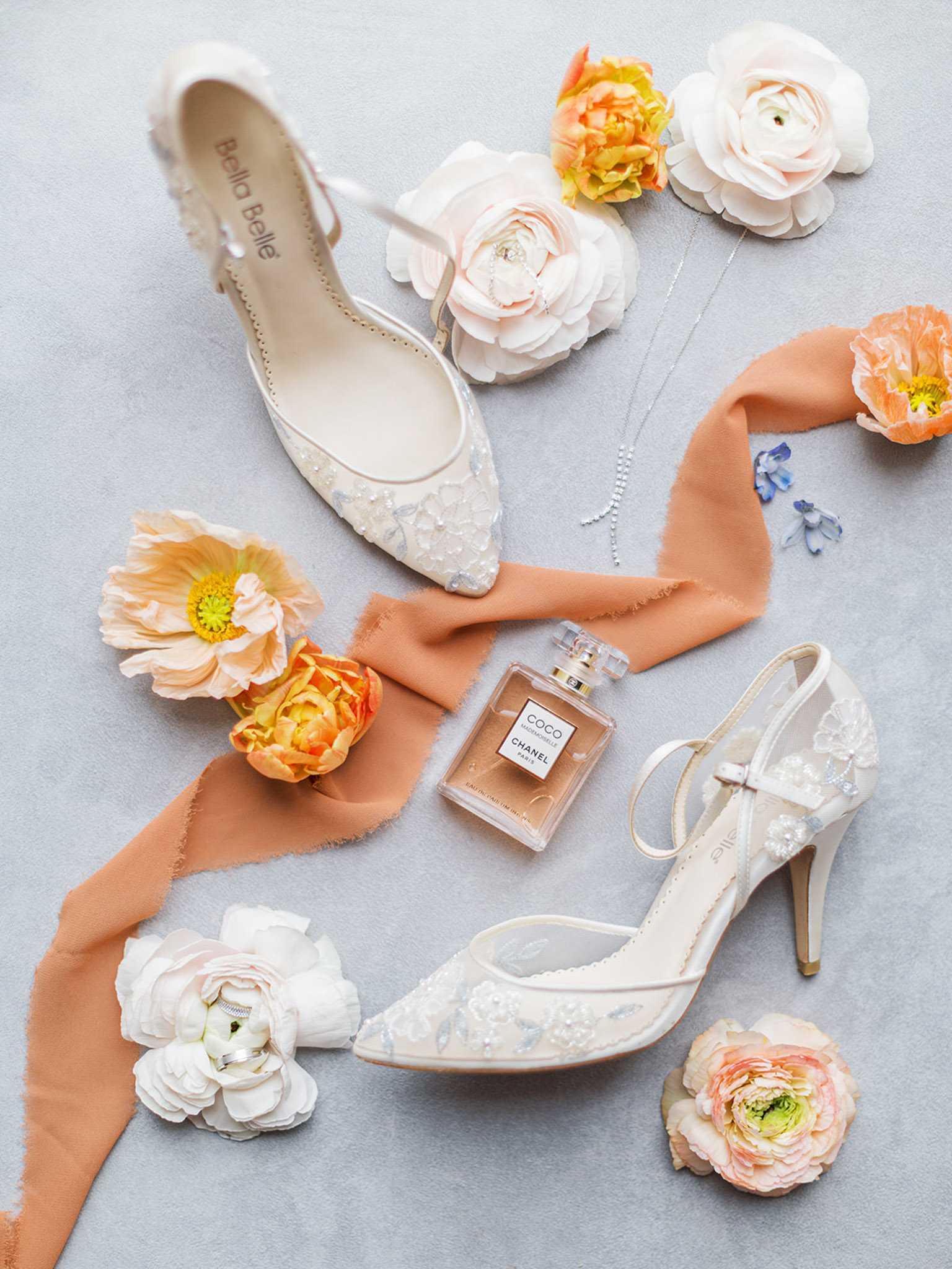 A flat lay detail shot of bridal accessories arranged on a light grey surface. Two pairs of Bella Belle ivory bridal shoes are featured — one embellished pointed-toe flat and one strappy heel with floral lace and beaded detailing — surrounded by loose blooms including blush and white ranunculus, peach poppies, orange peonies, and scattered blue delphinium petals. A terracotta silk ribbon weaves through the composition alongside a bottle of Coco Mademoiselle by Chanel perfume, a delicate silver chain necklace, and a diamond ring tucked into one of the ranunculus blooms. The overall color palette combines ivory, blush, peach, and terracotta with soft neutral tones.