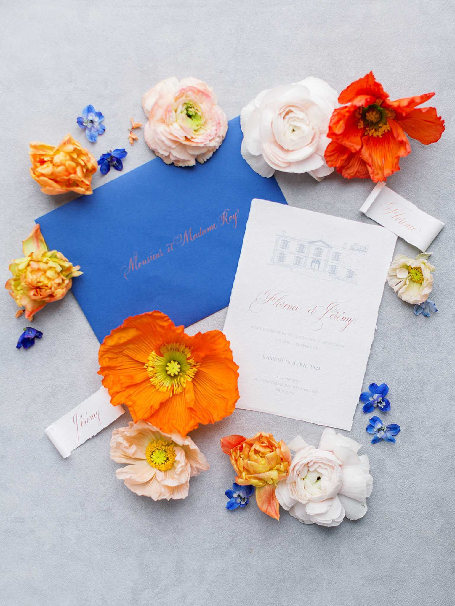 A flat lay detail shot of a French wedding stationery suite arranged on a light grey surface. The suite includes a cobalt blue envelope addressed in coral/terracotta calligraphy script reading 'Monsieur et Madame Roy,' a white deckle-edged invitation card featuring a hand-drawn illustration of a château at the top with the couple's names 'Florence et Jérémy' in coral script and event details printed below in French, and two small white ribbon place cards also written in coral calligraphy. The stationery is surrounded by loose flower heads including orange and blush Iceland poppies, blush and white ranunculus, orange garden roses, and small cobalt blue delphinium florets, creating a vibrant color palette of cobalt blue, coral, orange, and blush pink.