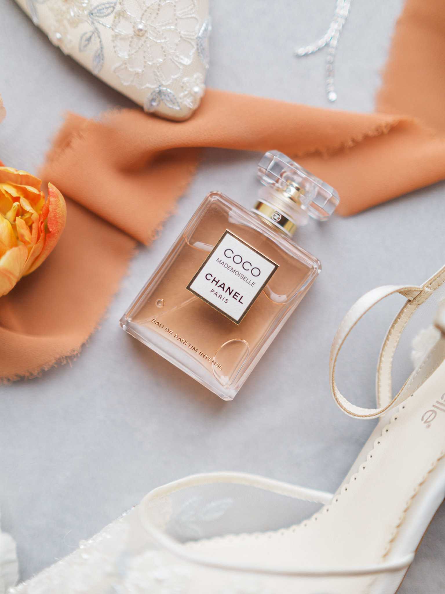 A bridal flat lay detail shot arranged on a light grey surface, featuring a bottle of Chanel Coco Mademoiselle Eau de Parfum Intense as the central element. Surrounding it are ivory lace-embroidered bridal shoes with satin ribbon ties, a terracotta-peach silk or chiffon ribbon draped loosely across the composition, a single orange ranunculus bloom in the upper left, and what appears to be a delicate crystal or silver jewelry piece in the upper right corner. The overall styling palette is warm peach, ivory, and gold, suggesting a classic bridal aesthetic.