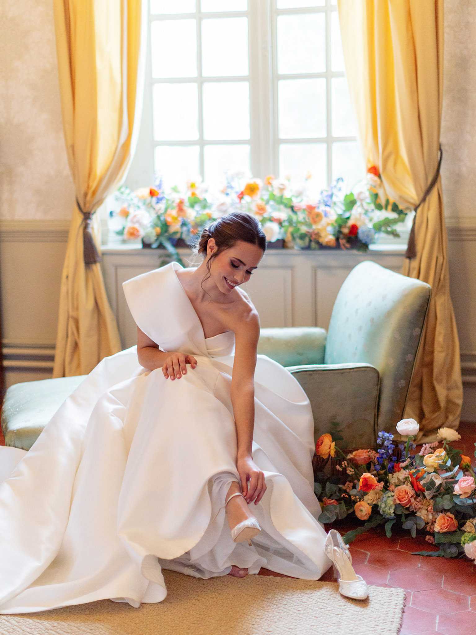 A bride is photographed getting ready indoors, seated on the floor beside a sage green upholstered armchair as she fastens a white heeled shoe onto her foot. She wears a structured ivory satin ball gown with a single oversized bow detail on one shoulder and a full skirt with a train. Her dark hair is styled in a low updo. Behind her, a tall window with gold satin drape curtains is lined with a low floral arrangement featuring peach, orange, coral, and blue blooms with greenery. A second loose floral cluster of similar colors — orange ranunculus, peach garden roses, blue delphinium, and eucalyptus — sits on the terracotta tile floor beside the chair. One white lace-trimmed heel rests on a woven rug in the foreground. The overall decor palette combines warm gold, sage green, and vivid multicolor florals in a classic French interior setting. This is a full-length portrait shot with a slightly editorial composition.